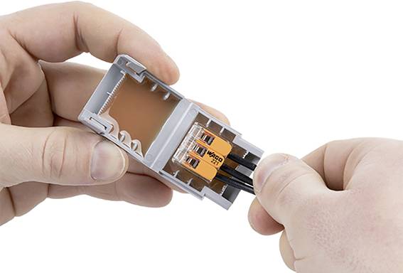 A person connects wires in a grey WAGO terminal block. The left hand holds the block open, while the right hand inserts wires.