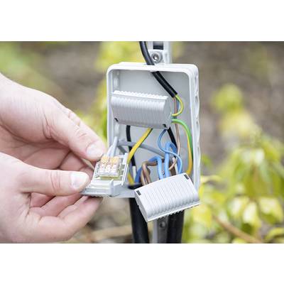 A person is installing electrical terminals in a grey junction box outdoors. Colourful wires are connected, with the surrounding environment blurred.