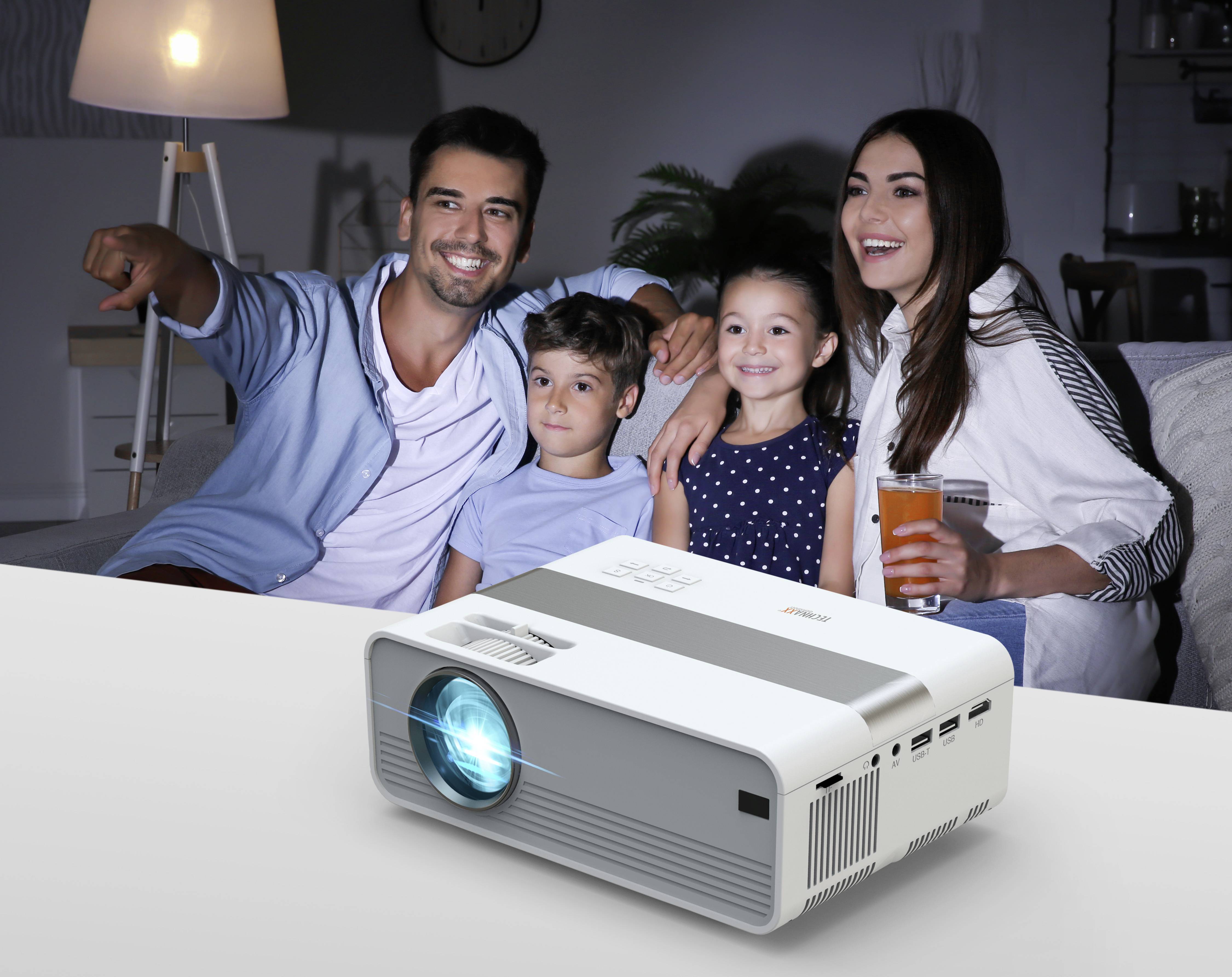A family with two children sits smiling on a sofa watching a film. A projector on the table emits a blue light.