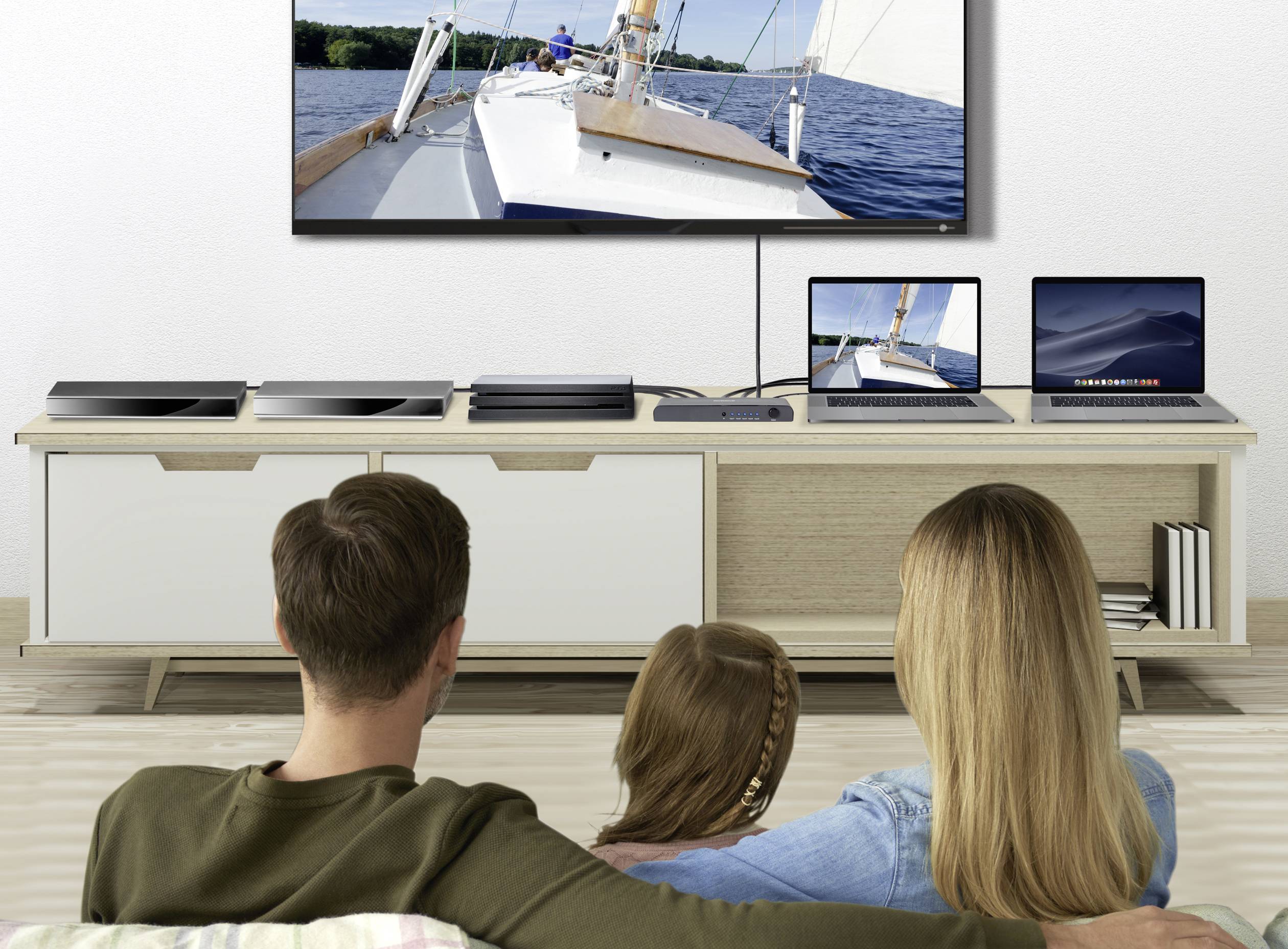 A family is sitting on a sofa, looking at a large television showing a sailing boat on the water.