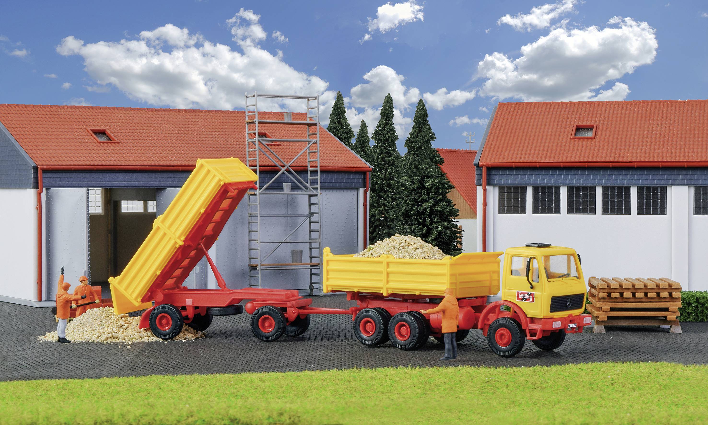 A lorry is tipping gravel onto a yard in front of storage buildings. Two workers are standing nearby, and a stack of wooden planks lies close by.