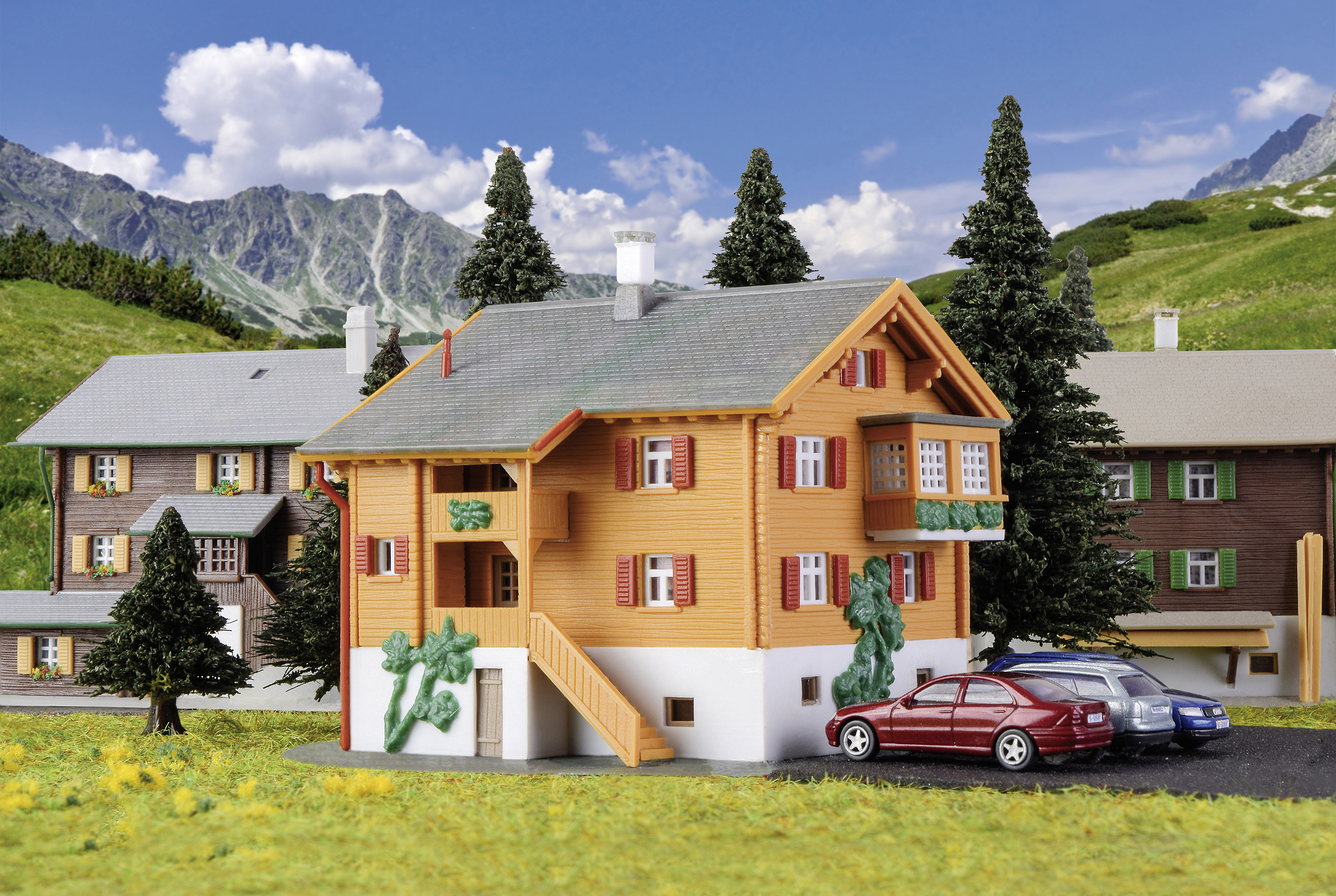 A colourful model house in an alpine landscape, surrounded by trees and neighbouring houses. Two cars are parked beside it. Mountain backdrop in the background.