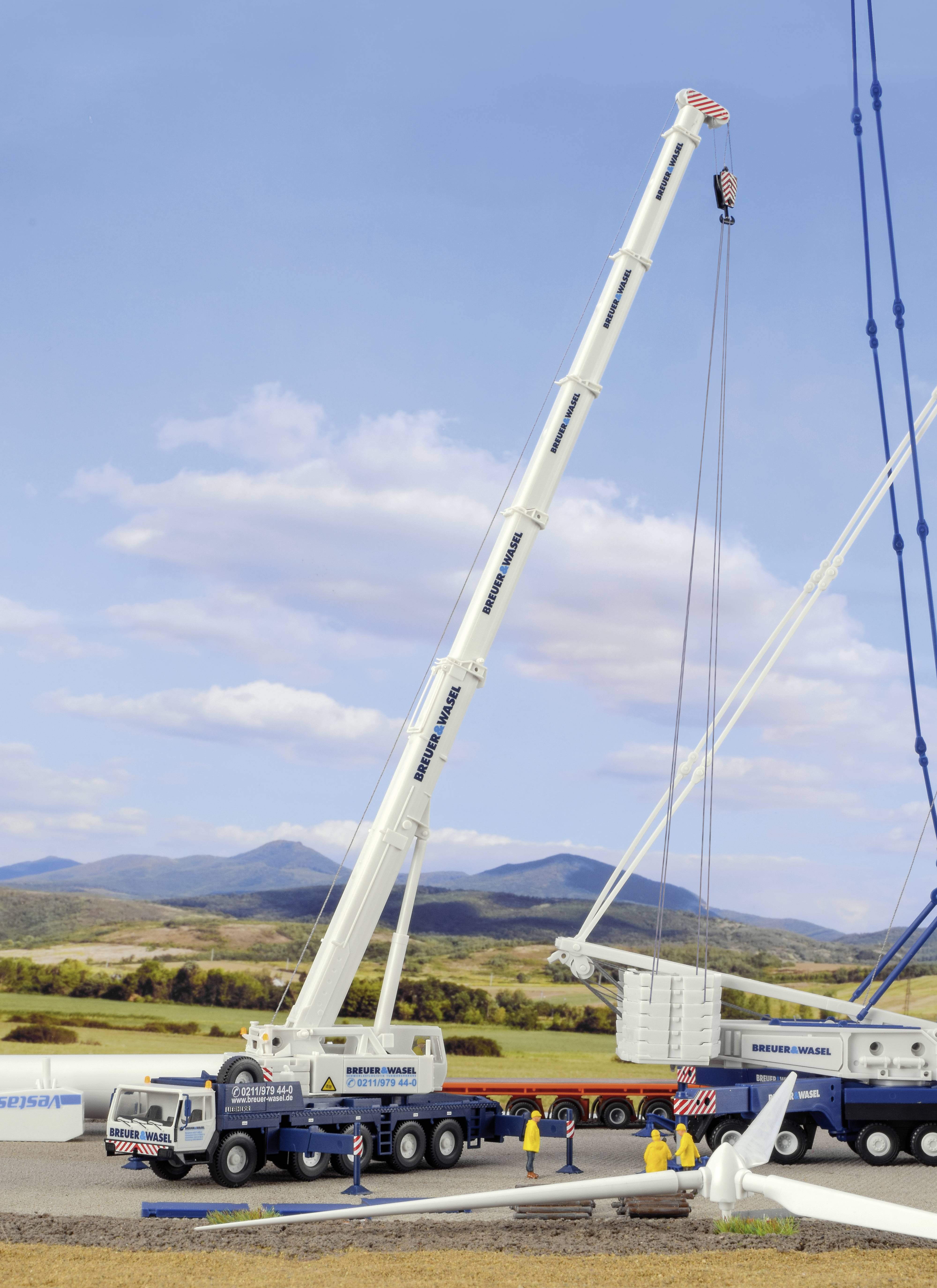 A large crane lifts a heavy structural component at a construction site in a rural area. People wearing safety clothing observe the procedure.