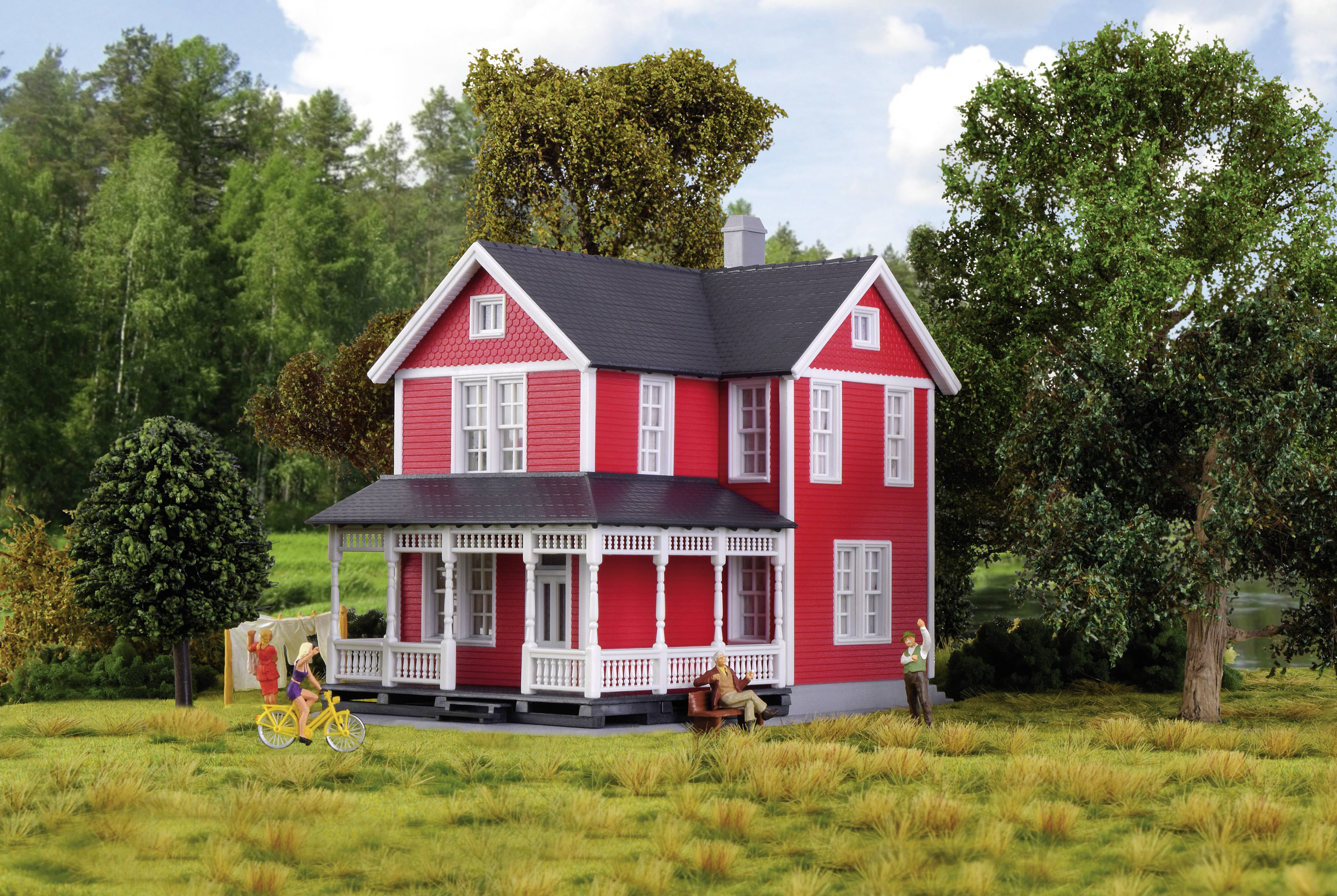 A red house with a veranda stands on a grassy field, surrounded by trees. One person is cycling, another is sitting in front of the house.