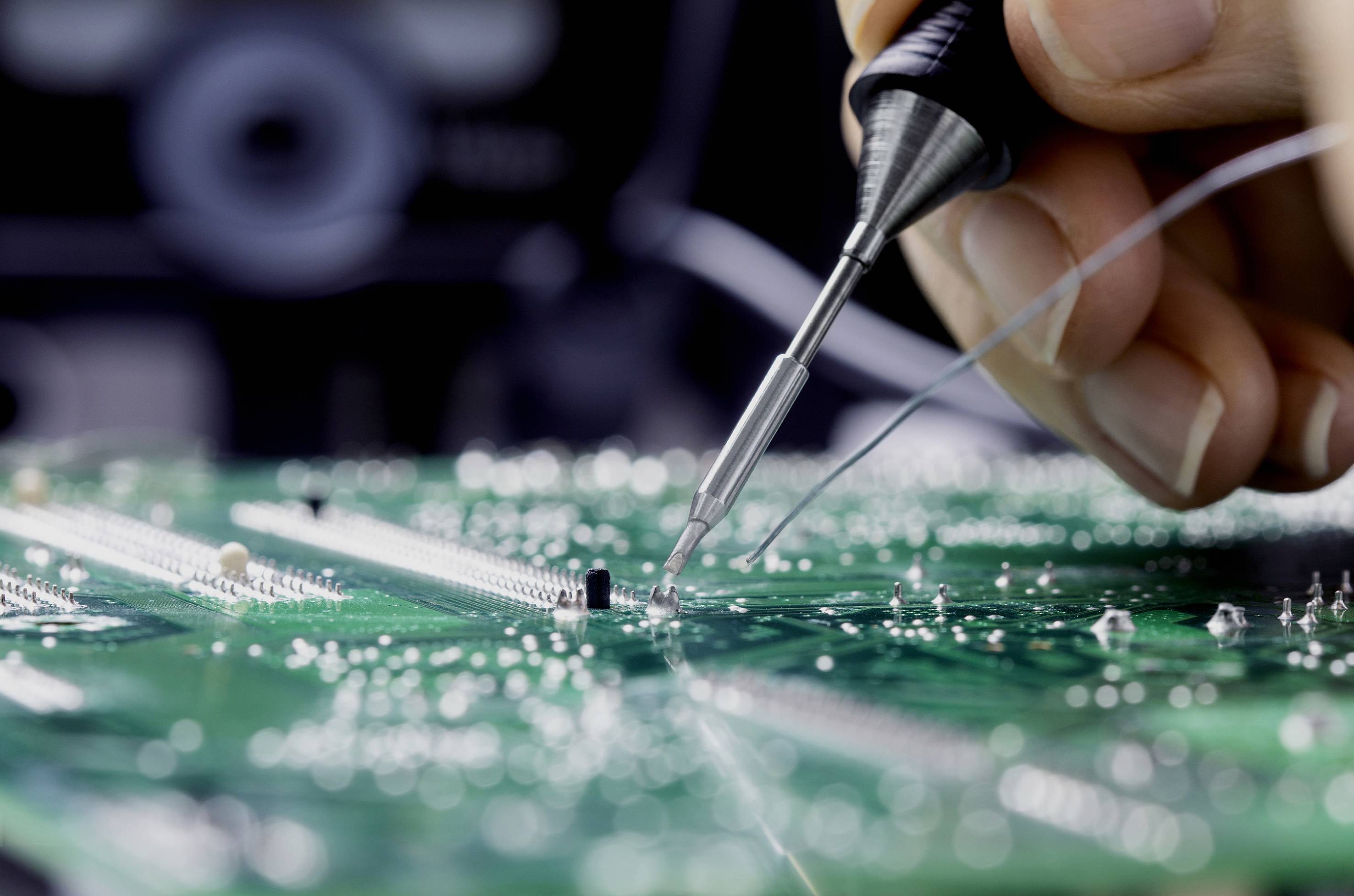Close-up of a person working with a soldering iron on a green circuit board. One hand is holding the iron to connect components.