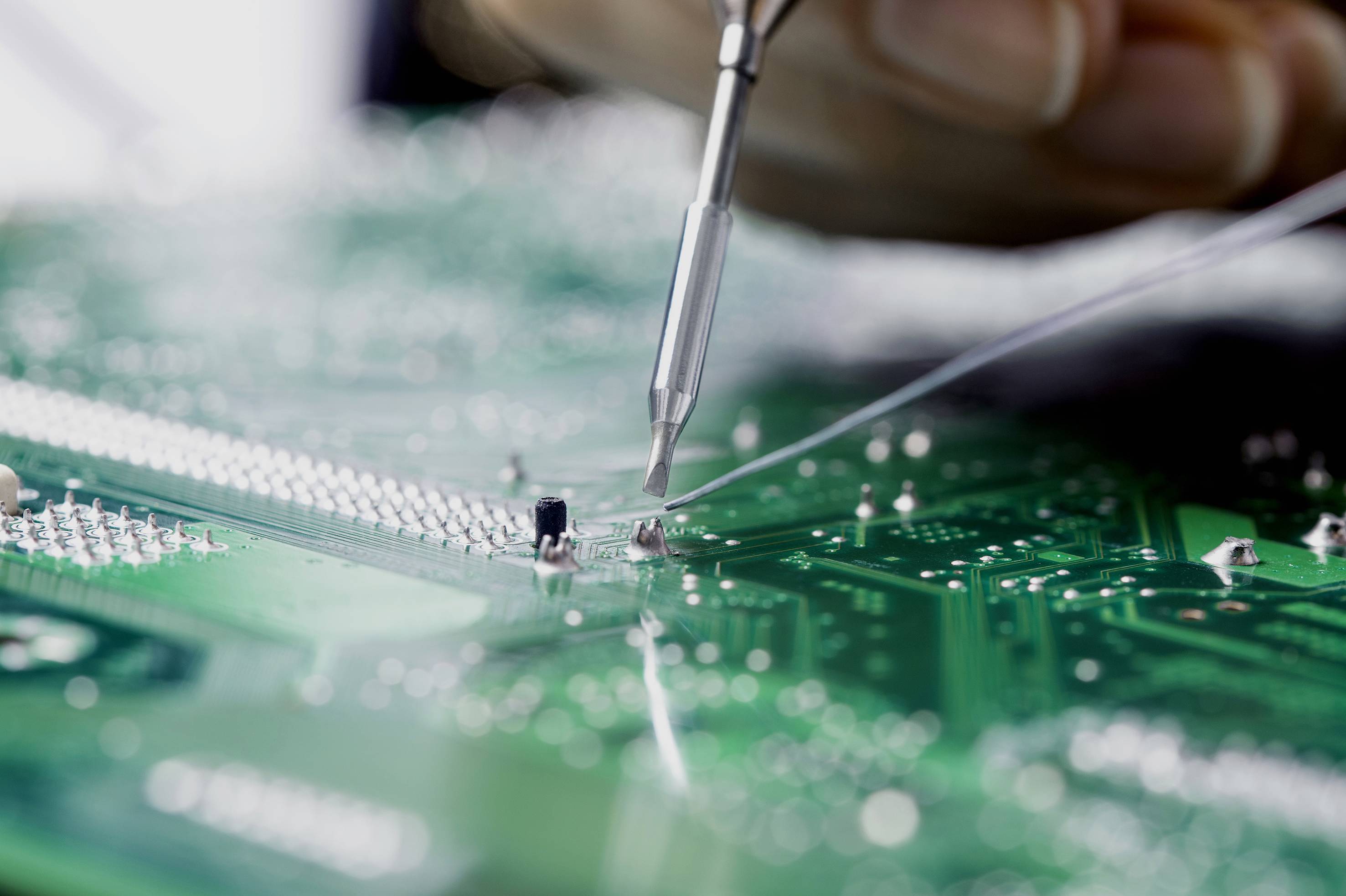 Close-up of a circuit board, with a hand using a tool to solder or repair electronic components.