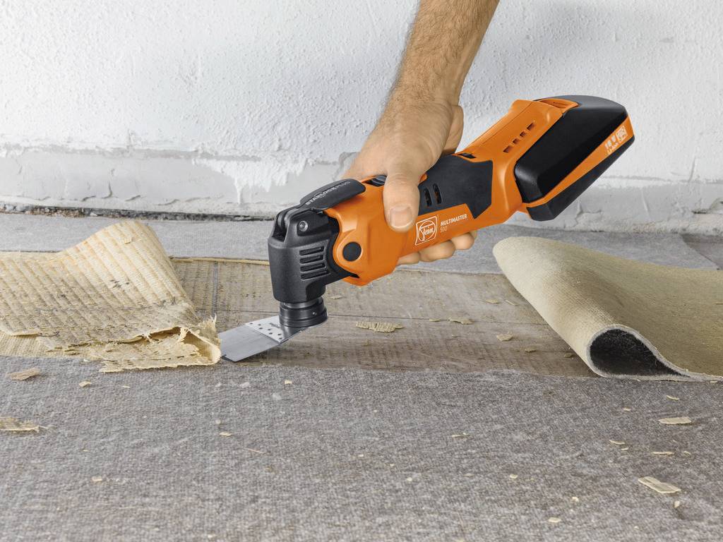A hand is holding an orange multi-functional tool that is removing old carpet and floor covering from a concrete floor.