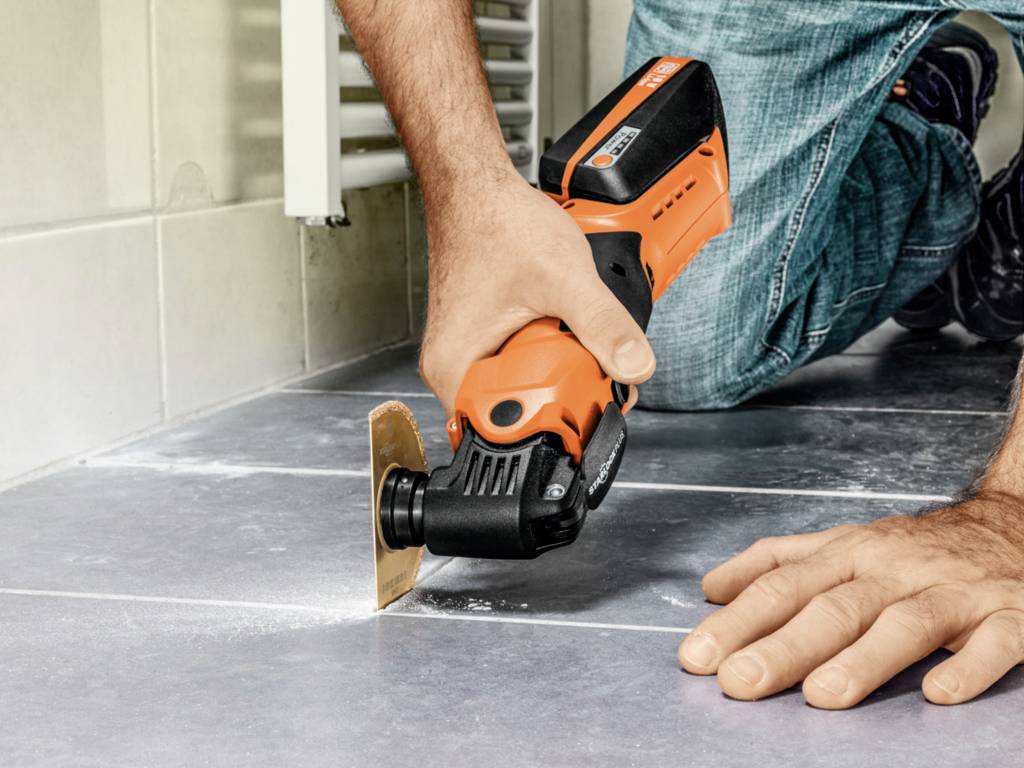 A person is using an oscillating tool to cut tiles in a bathroom. The tool has an orange handle.