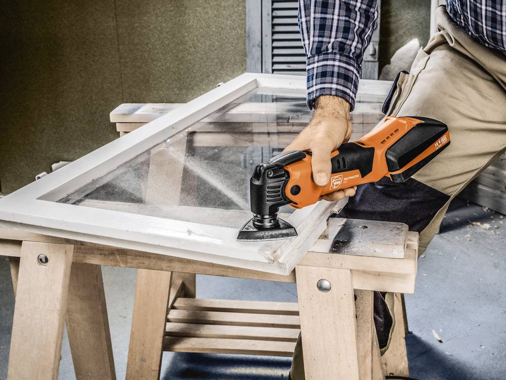 A person is working on a window frame with an electric multi-tool on a workbench.