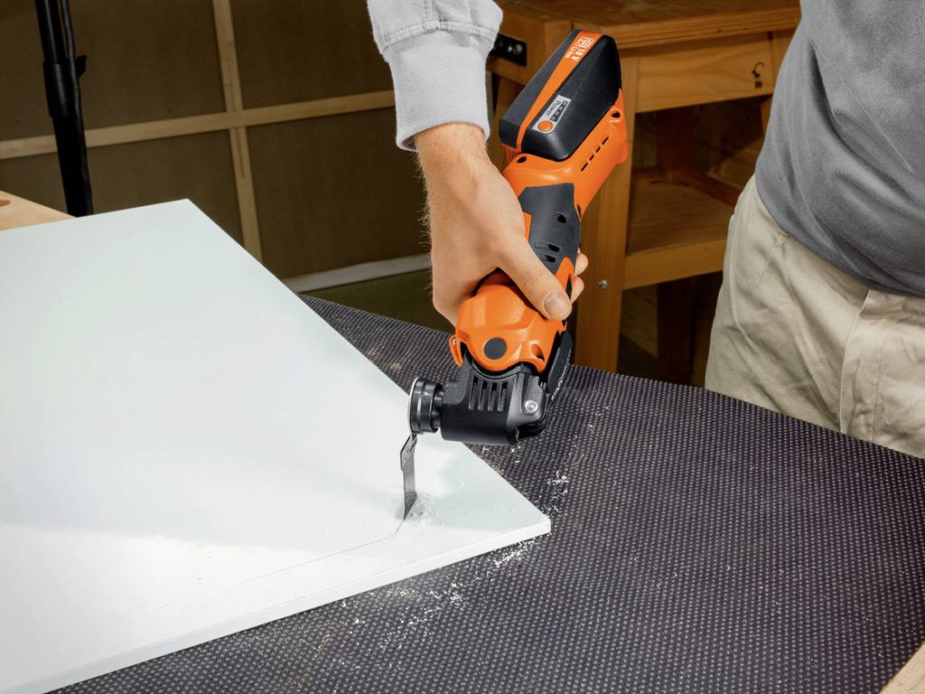 A person is cutting a white board with an orange oscillating multi-tool on a table.