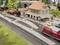 Model railway scene with a small station. People waiting on platforms. Trains on tracks. Buildings with red roofs in the background.