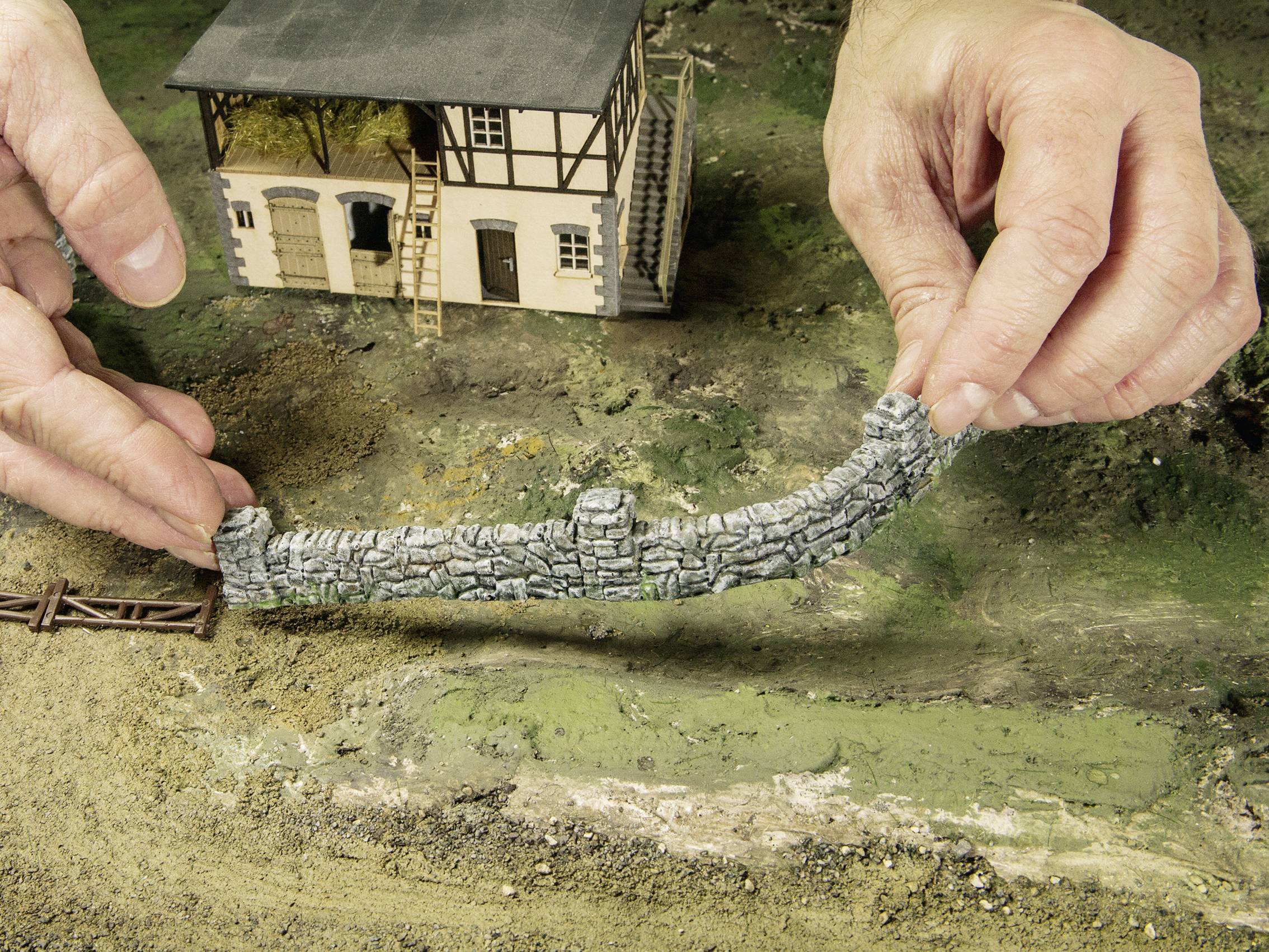 A person positions a small stone wall in front of a model house in a miniature landscape. Hands reach for the wall.