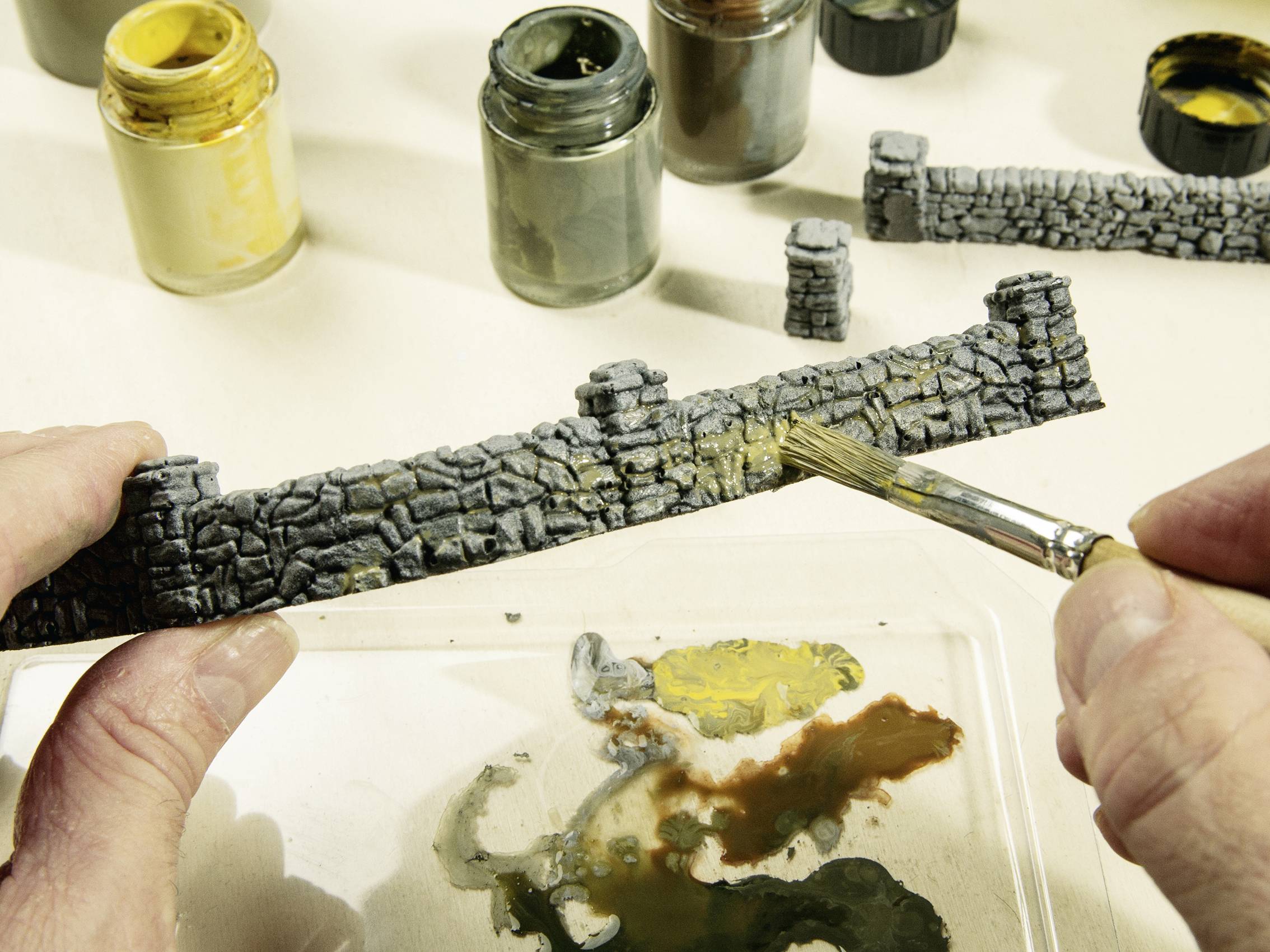 A person is painting a miniature wall section with grey paint. A brush is being used, with several paint pots and a colour palette visible in the background.