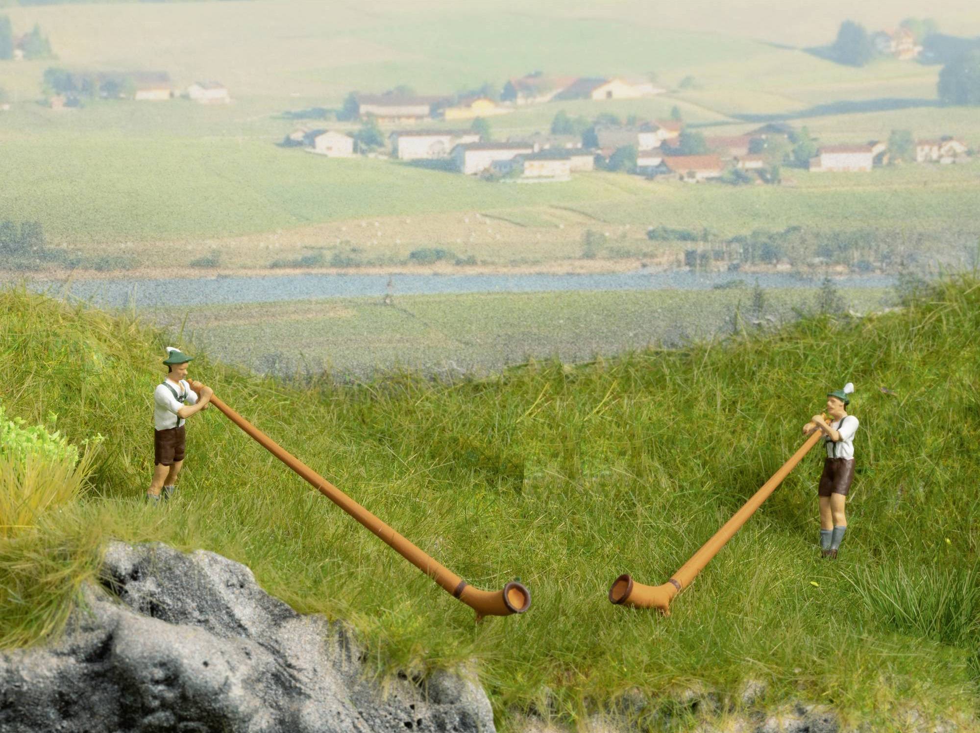 Two people in traditional clothing play alpine horns in a green landscape with a village in the background.