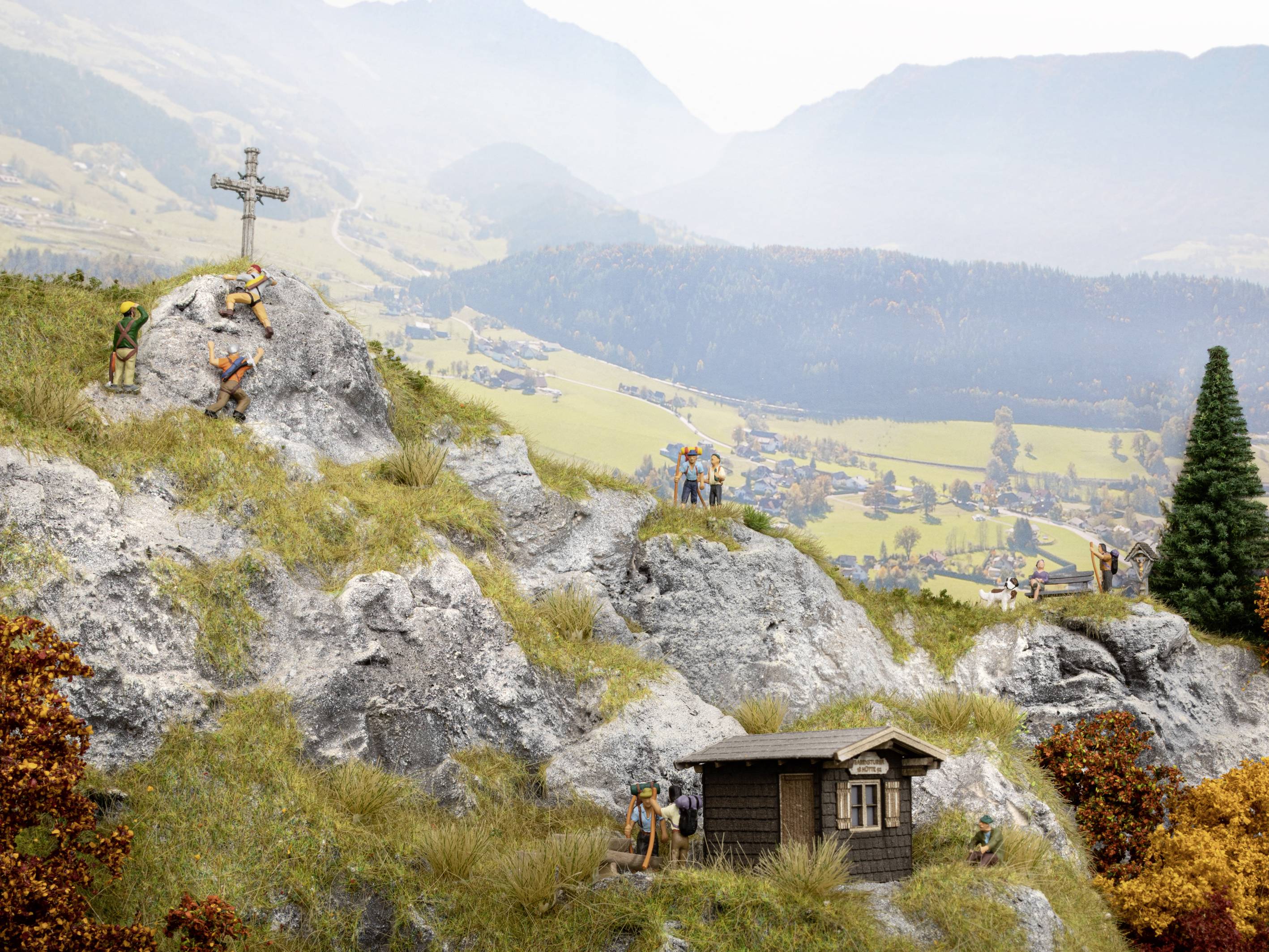 Miniature mountain landscape with climbers on rocks, a wooden cabin and a cross. Villages and valleys can be seen in the background.<br><br>Note: The translation is already in British English, so no further modifications were necessary.