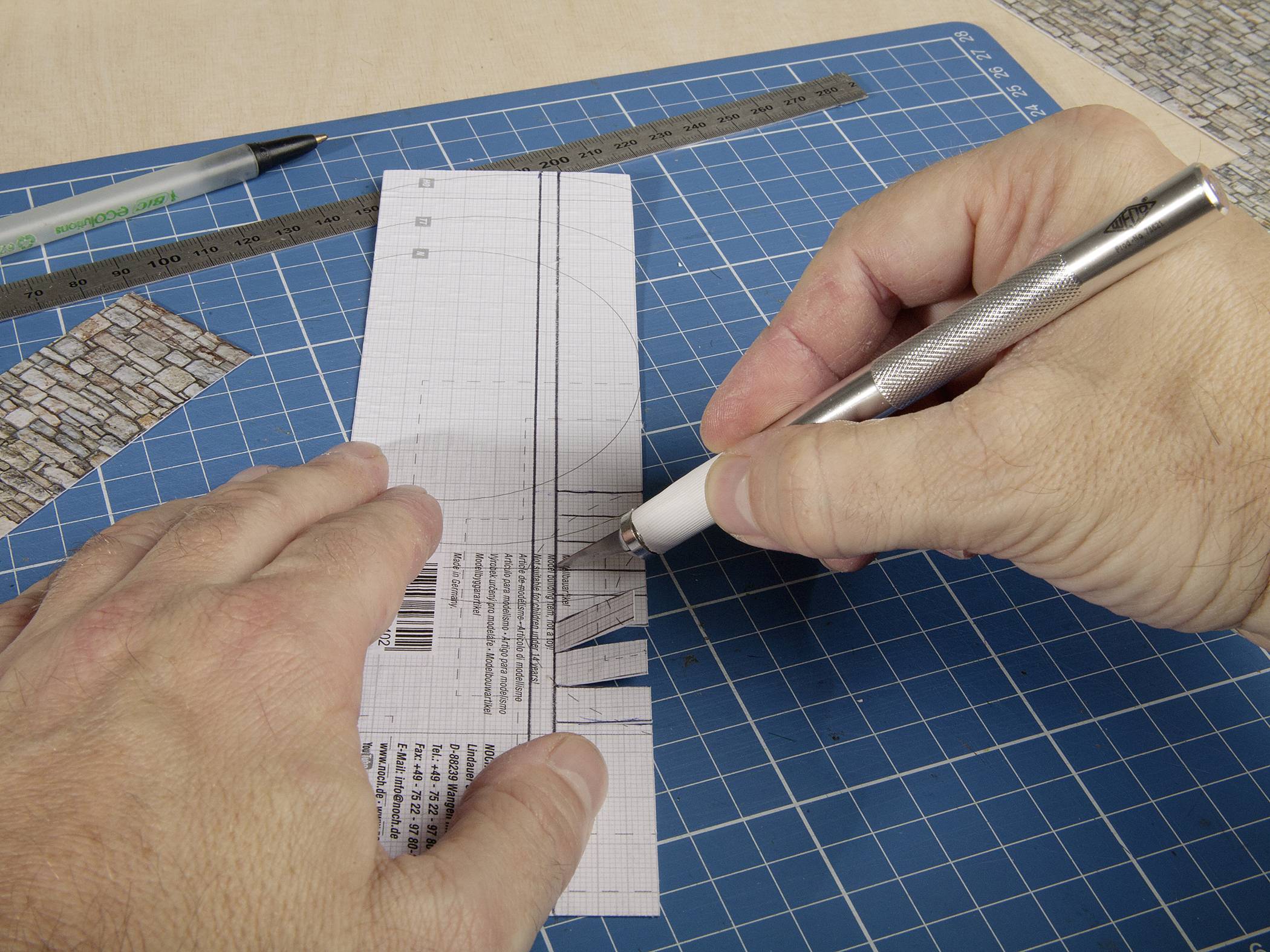 A person is cutting a rectangular piece of paper with a precision knife on a blue cutting mat, with a pen and ruler beside it.