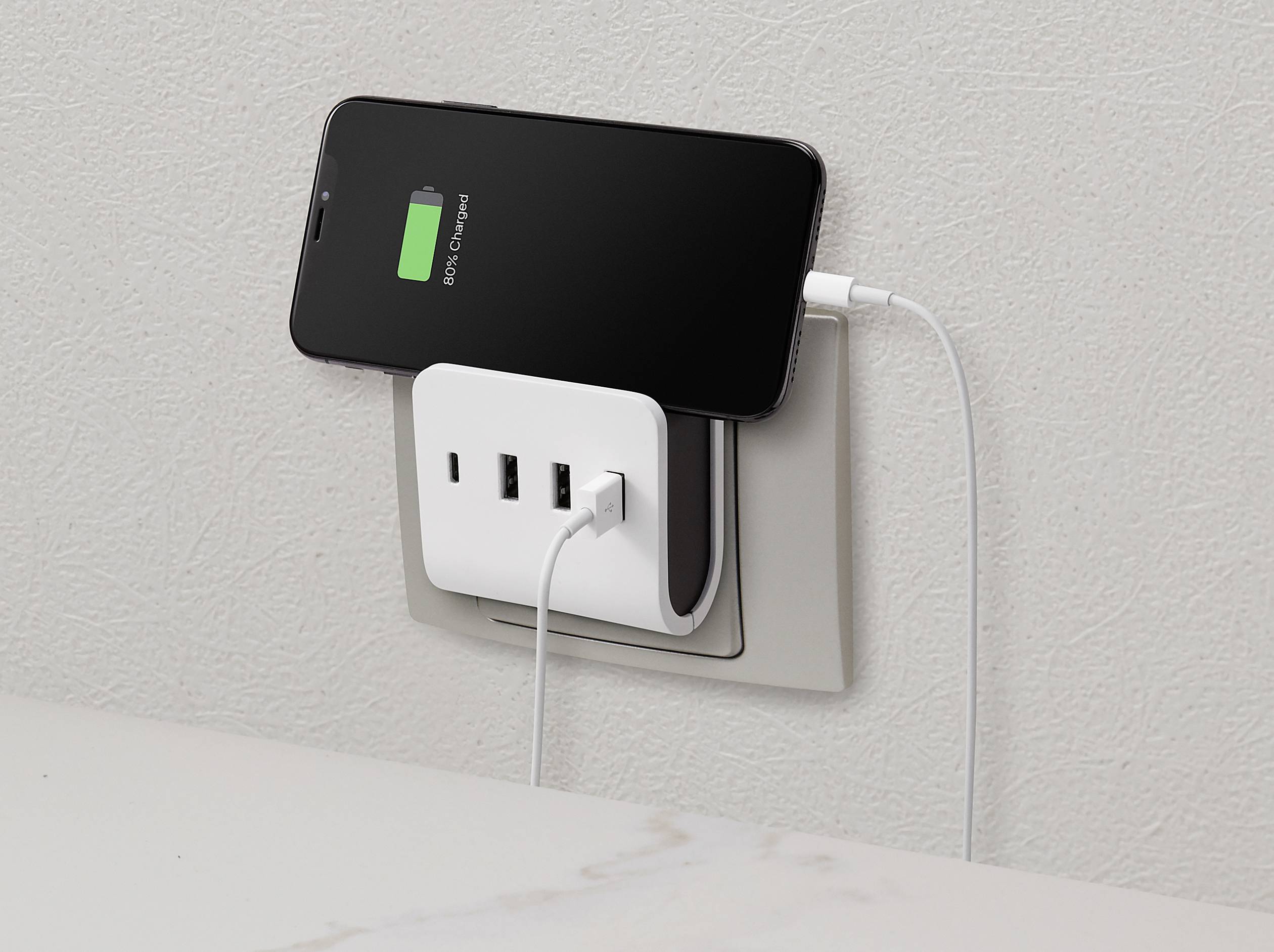 Smartphone charging at a wall socket. The battery displays a green charging indicator. Three USB ports available on the charger.
