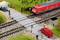'Model railway scene with level crossing: Red train crossing the road, red car waiting. Workers wearing safety helmets.'