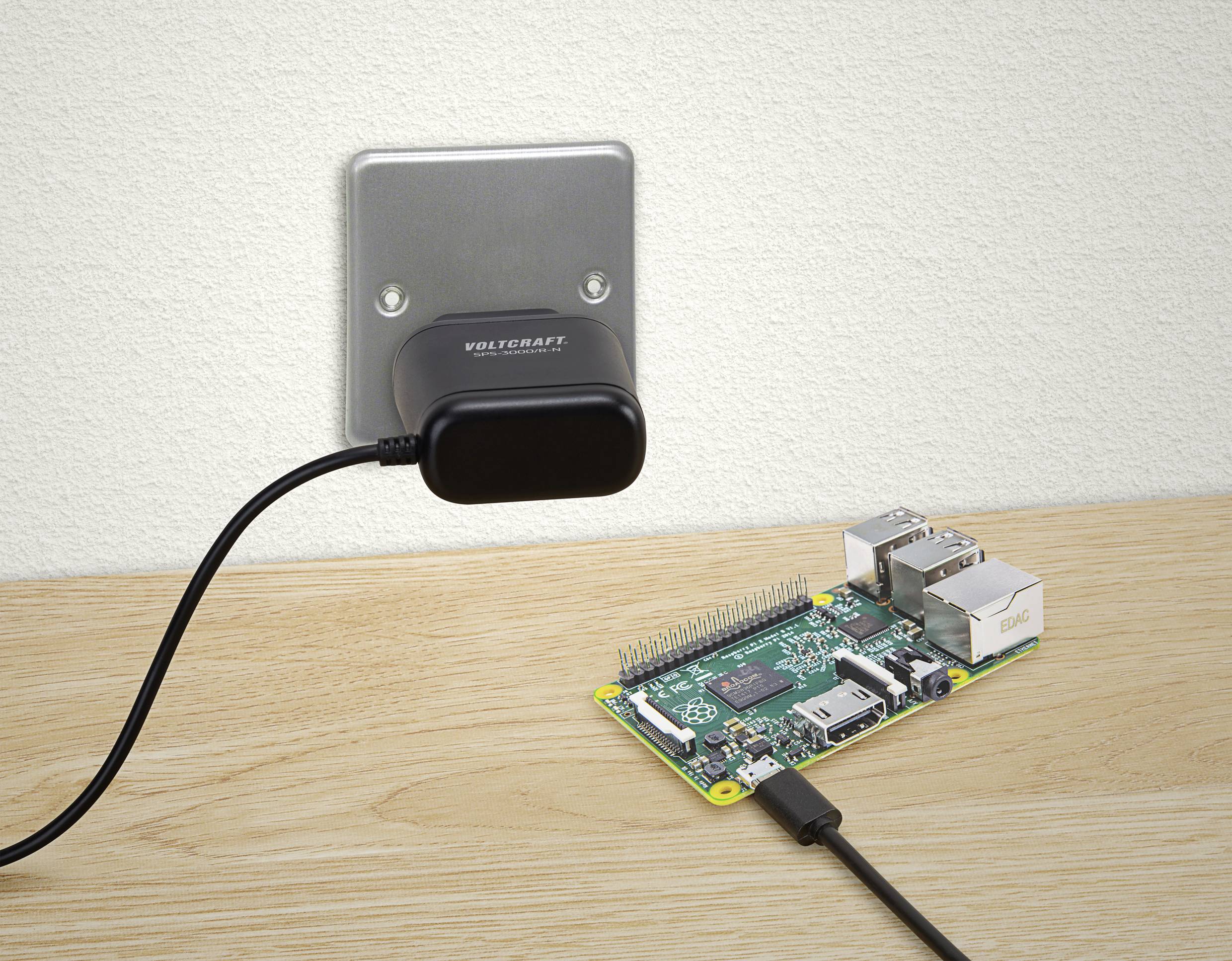 A small computer is connected to a wall-mounted power supply via a black cable. They are situated on a wooden table.