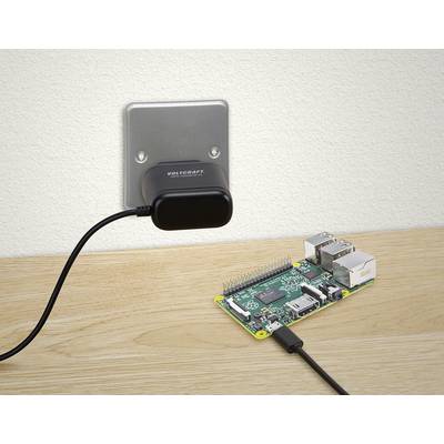 A small computer is connected to a wall-mounted power supply via a black cable. They are situated on a wooden table.