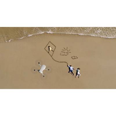 A couple lies on the beach, holding a drone drawn as a kite, with a child in the kite. Next to them, a sun and two clouds are drawn in the sand.