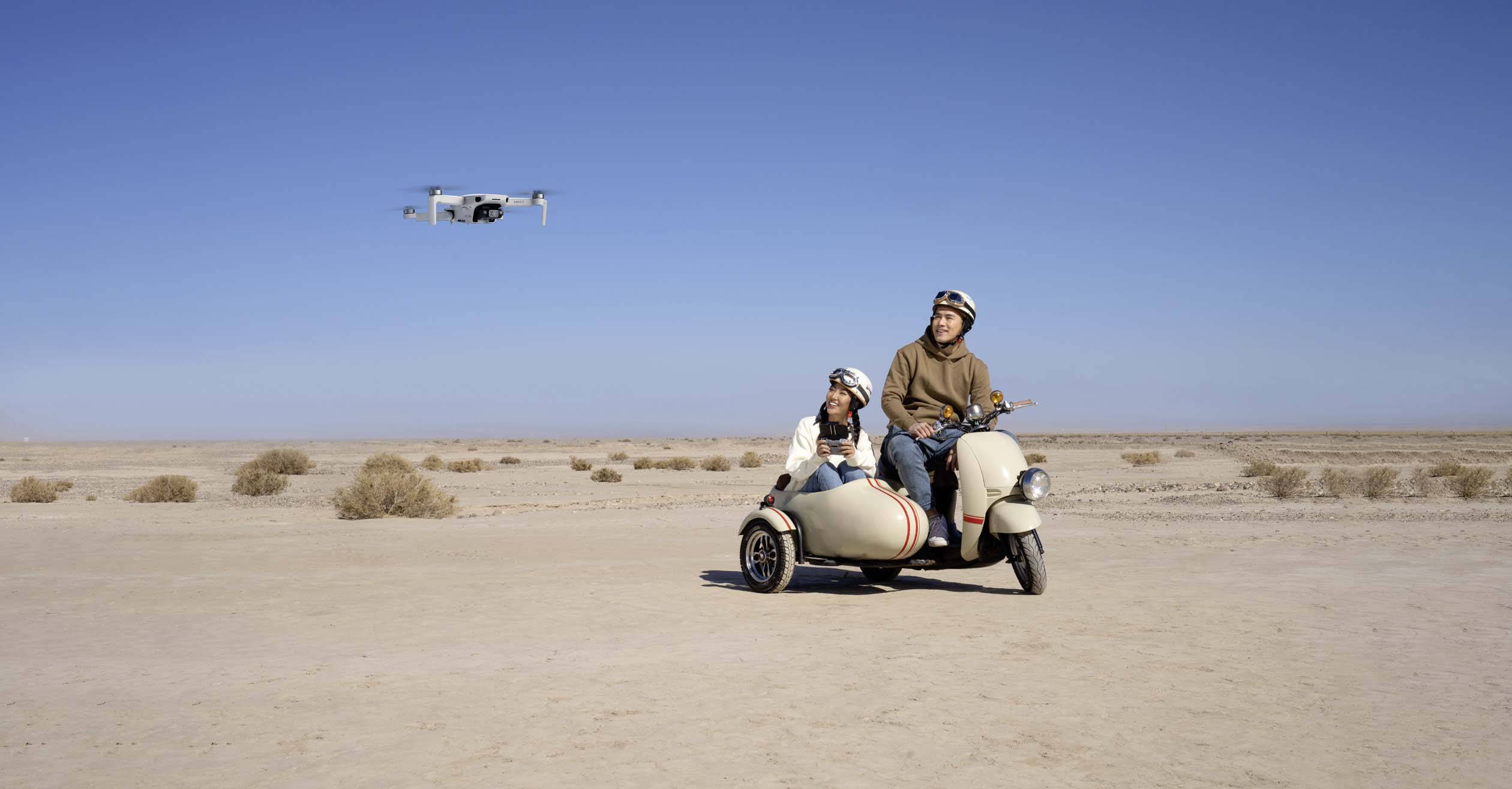 Two people are sitting on a motorcycle with a sidecar in a desert landscape; a drone is flying nearby in the blue sky.