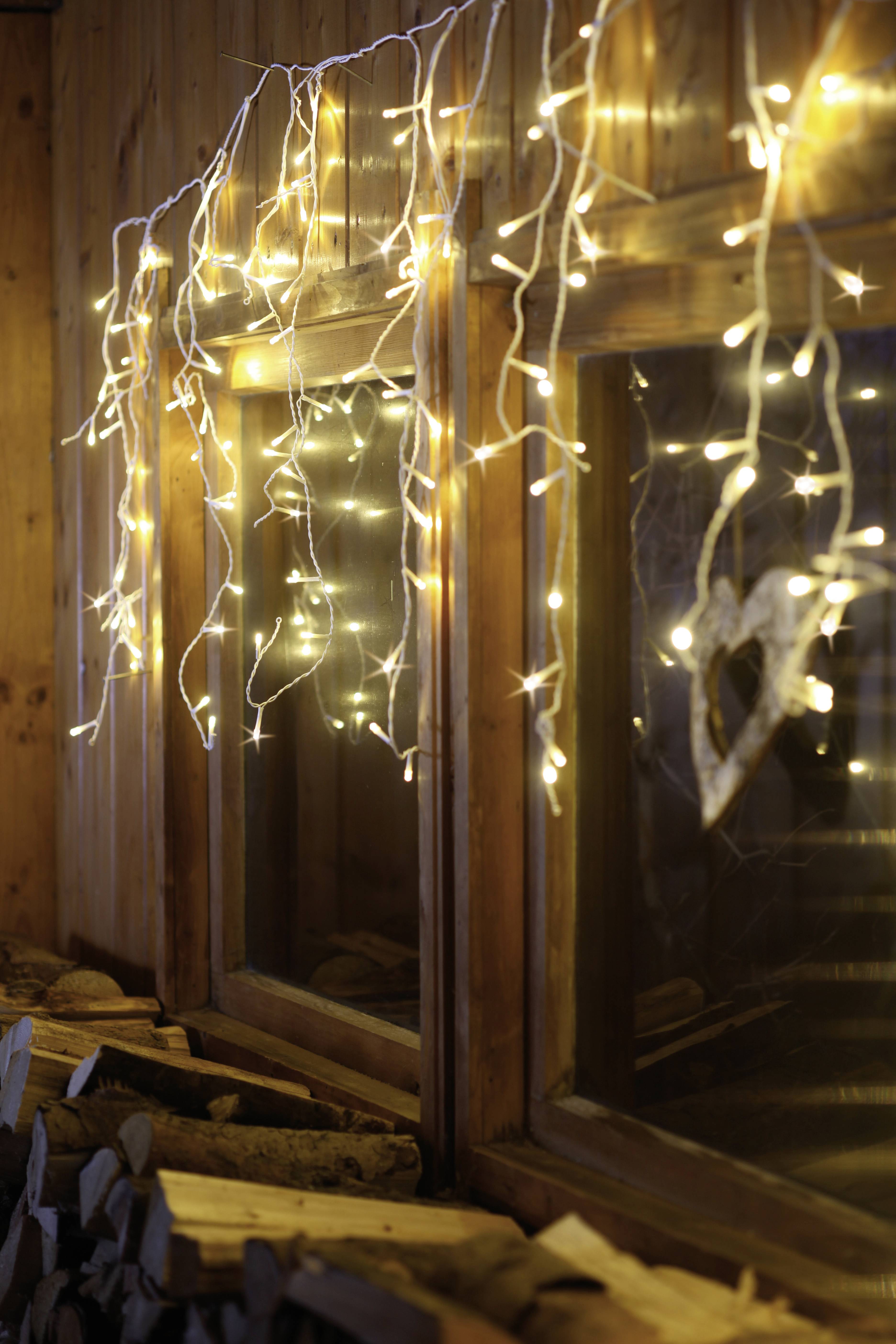 Fairy lights hang in front of a window, with stacked logs of wood beside them. The lighting creates a cosy, festive atmosphere.