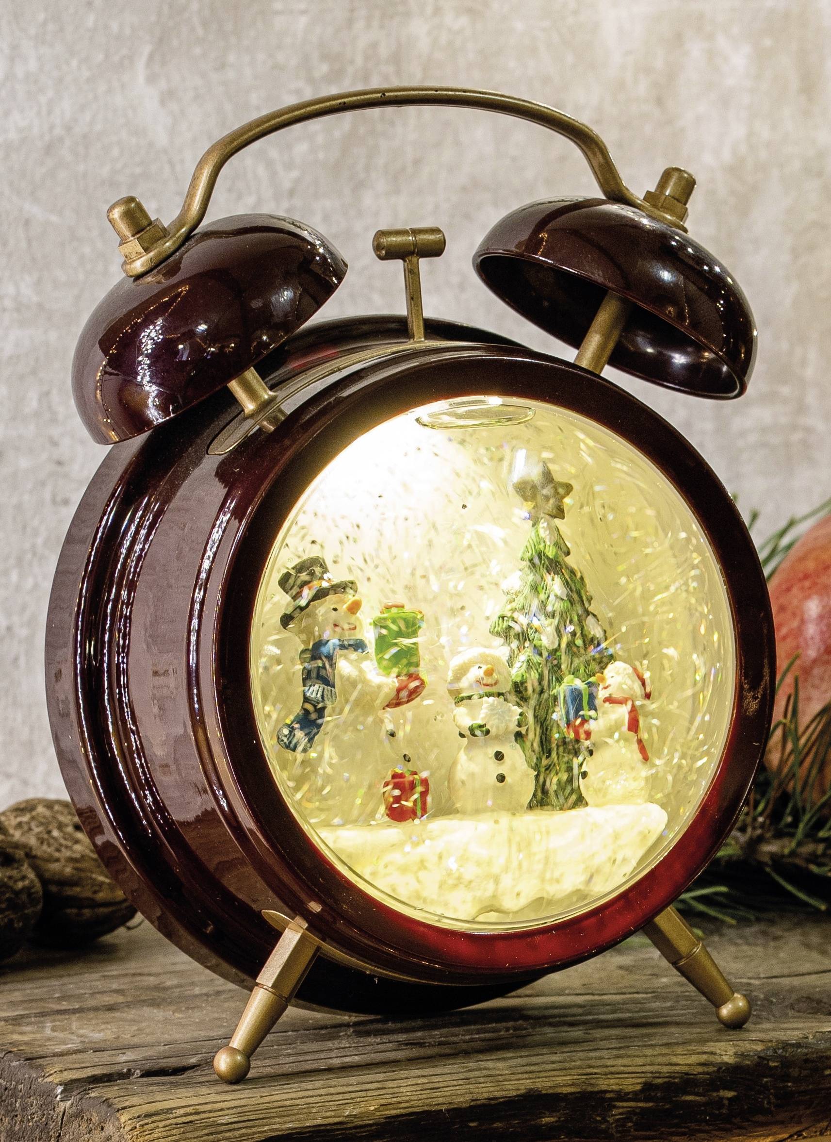 A decorative clock in the shape of an alarm clock depicts a wintry scene with three snowmen and a Christmas tree inside.