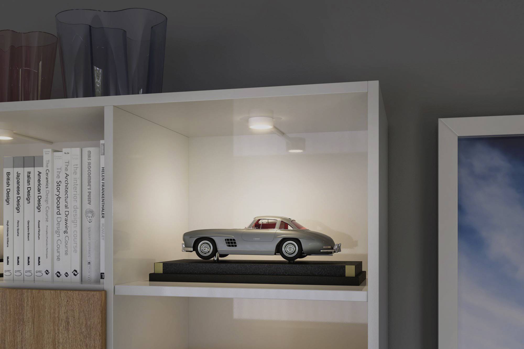 A silver model car sits on a shelf beside some books. It is illuminated from above and serves as a prominent decorative feature.