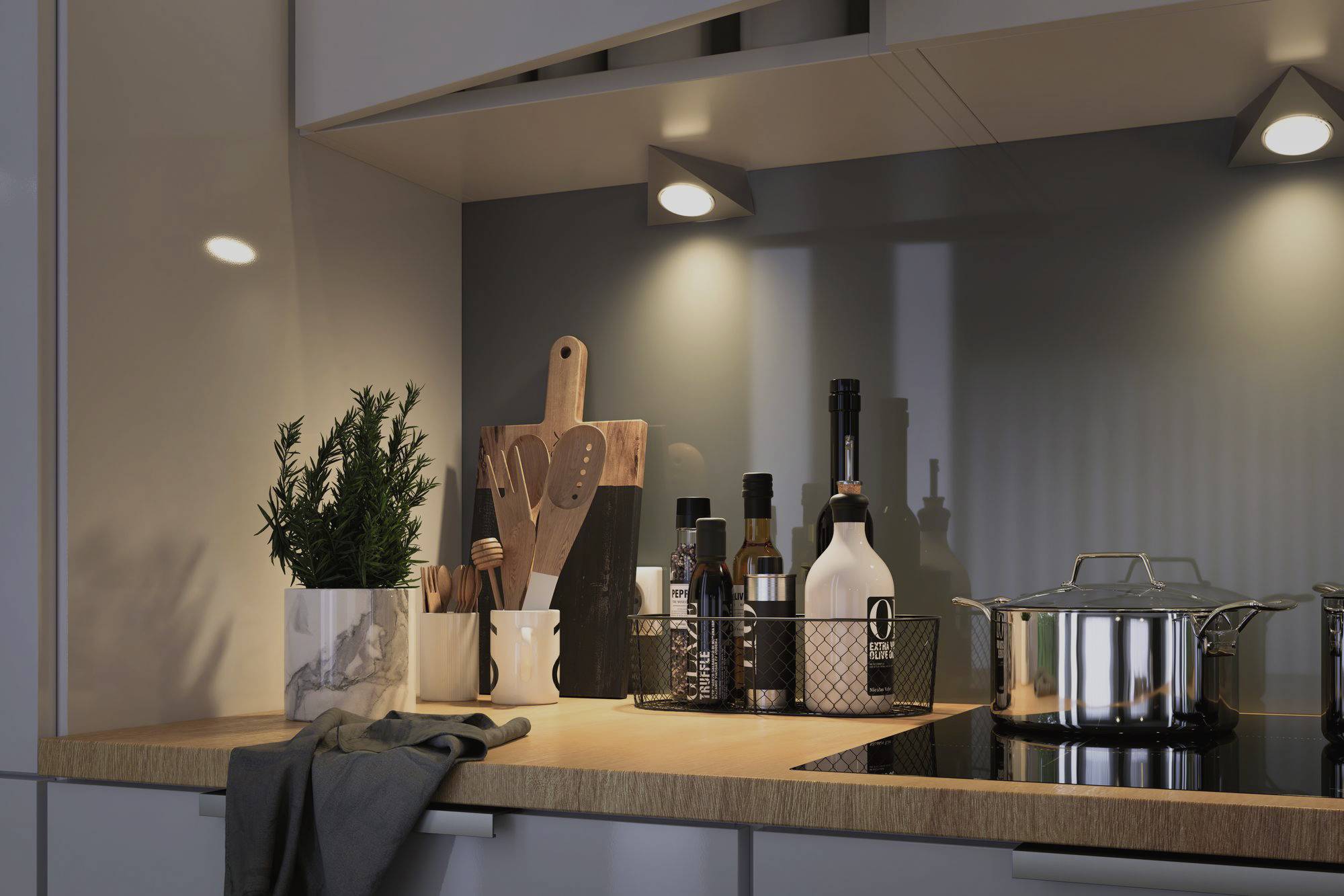 In a modern kitchen, plants, a chopping board and various bottles sit on the worktop next to a cooking pot on the hob.