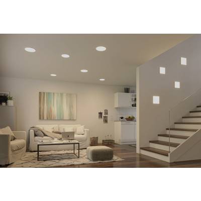 Bright living room with a white sofa and wall art. Modern lighting on the ceiling and stairway wall, with an open-plan kitchen in the background.