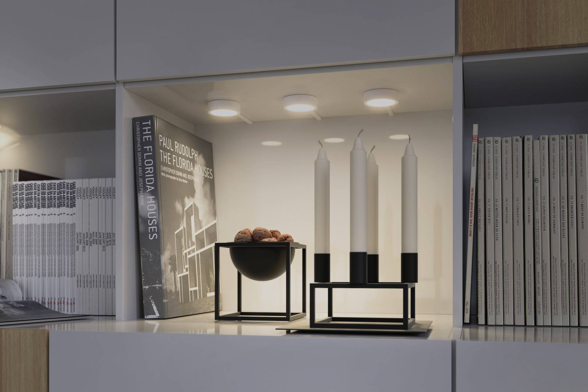 Bookshelf with books, four white candles in a black holder and a bowl of nuts. Warm light creates a cosy atmosphere.
