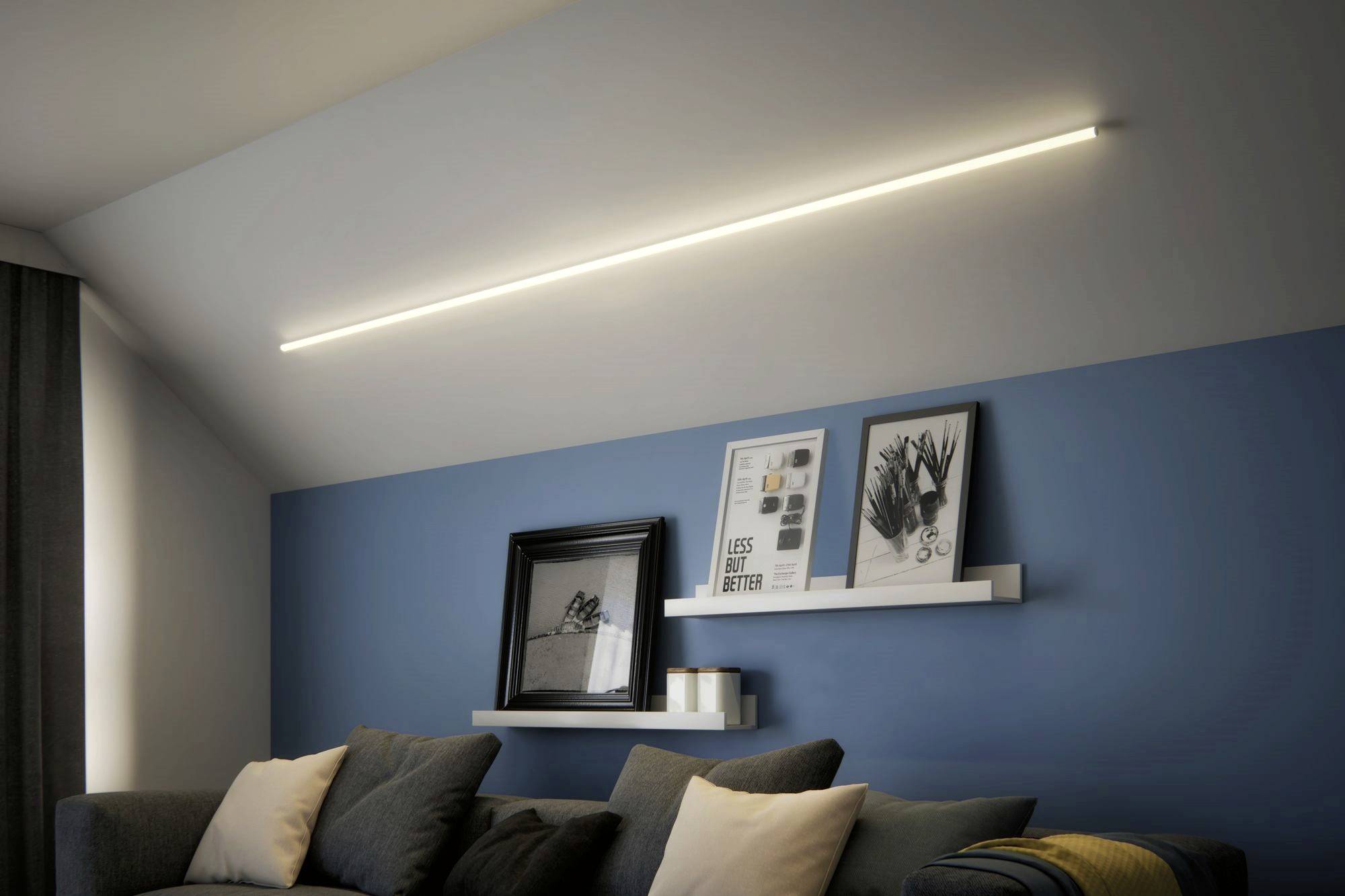 Living room with a blue wall, illuminated by a long, horizontal light fixture on the ceiling. Two shelves with pictures and candles above a grey sofa.