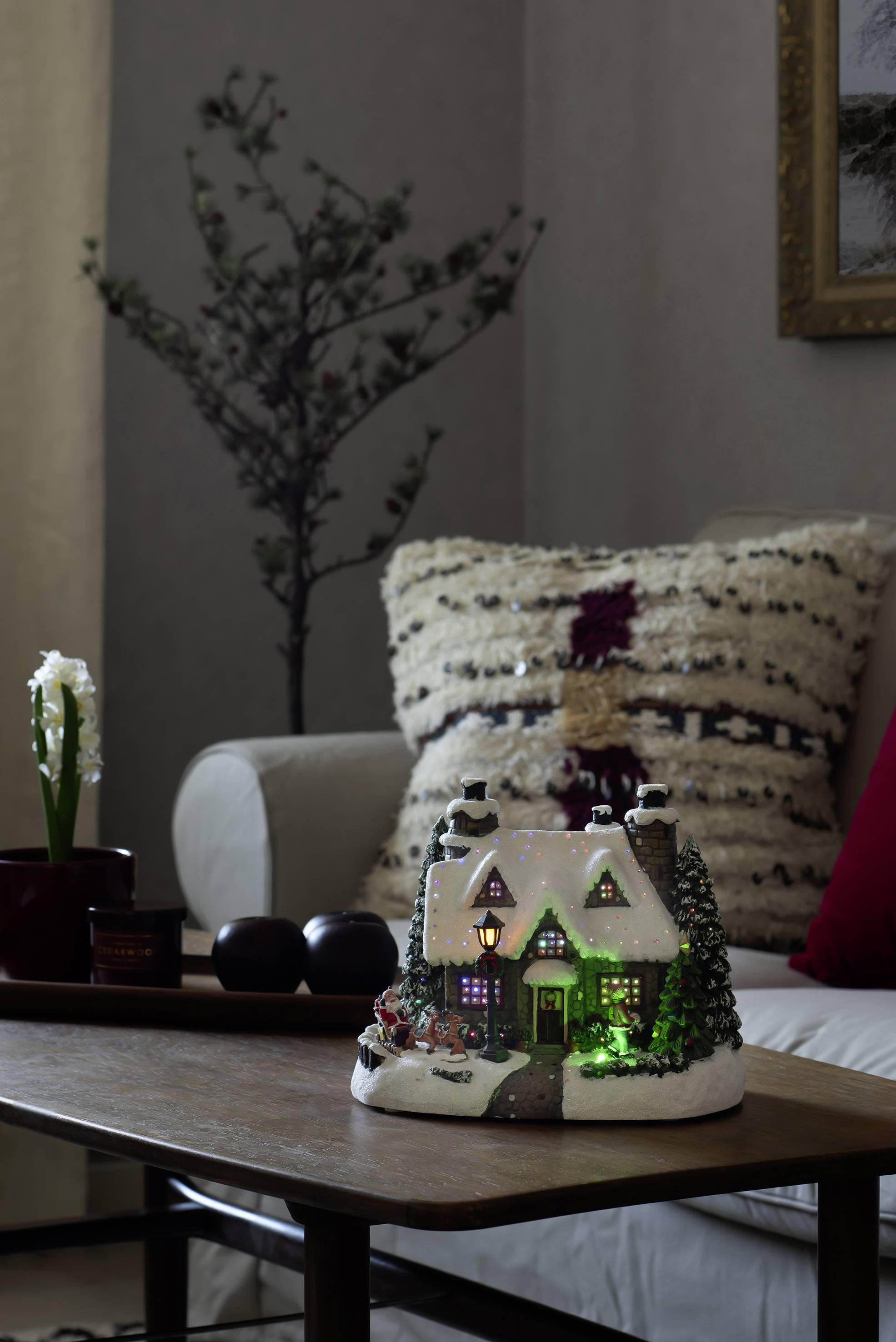 An illuminated, decorative little house sits on a table, flanked by candles and a small flower, in front of a sofa with cushions.