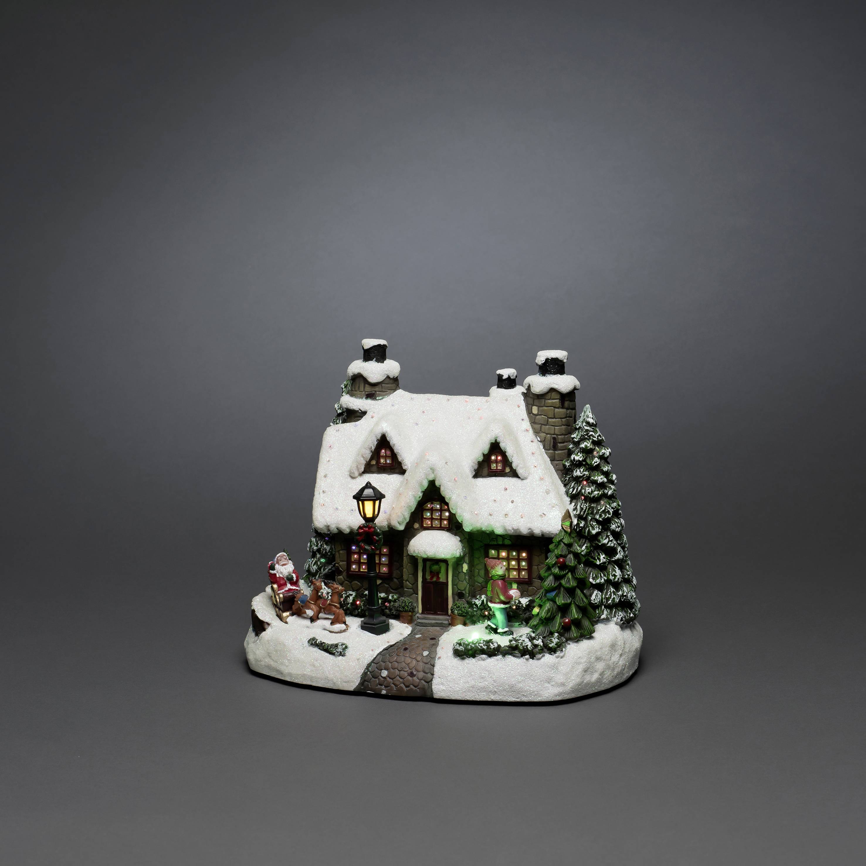 Small, illuminated Christmas village house with a snow-covered roof, Christmas tree, and Santa Claus figurine in the foreground against a grey background.