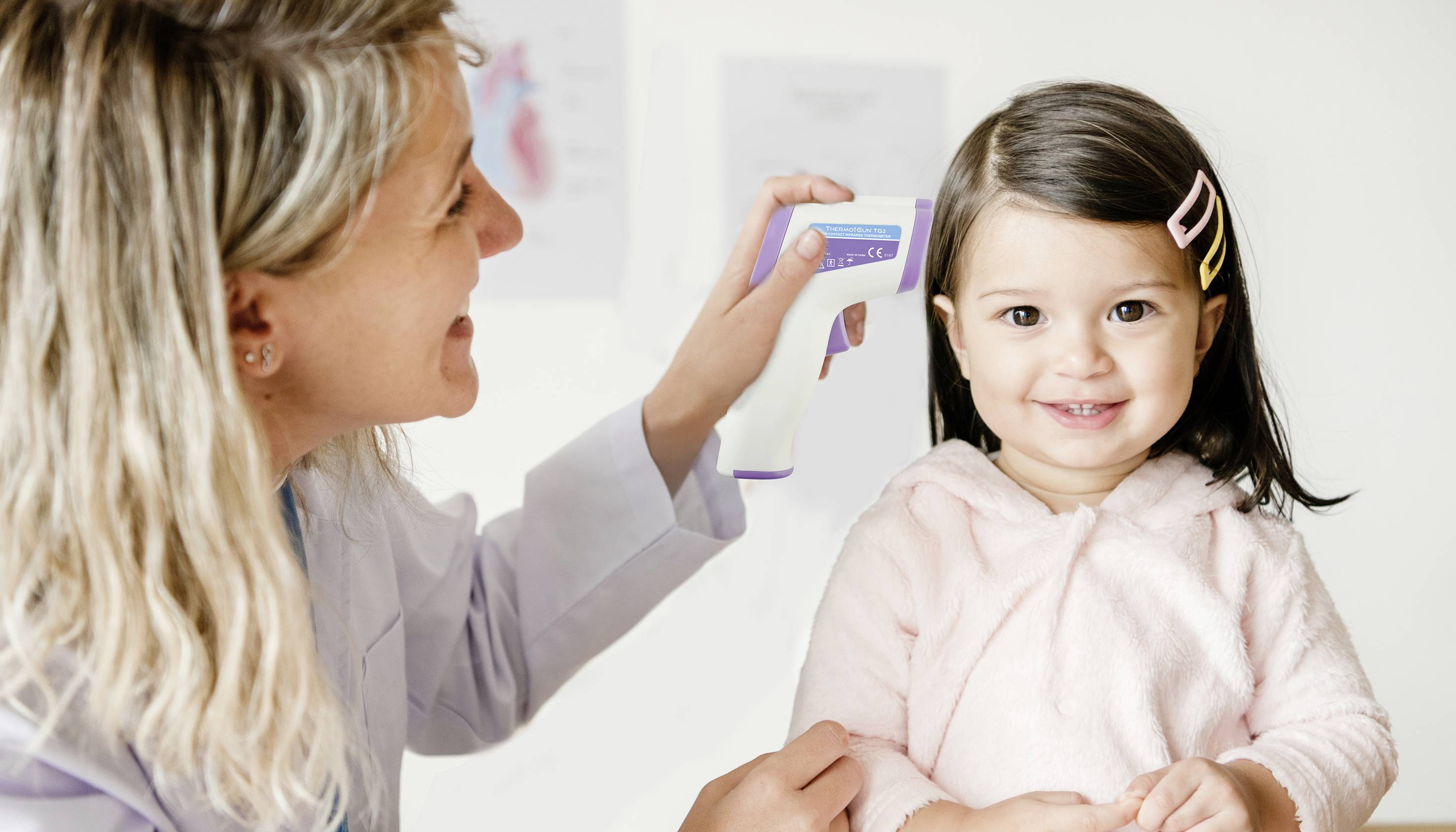 A woman is measuring the temperature of a smiling little girl in pink clothing using a non-contact thermometer.