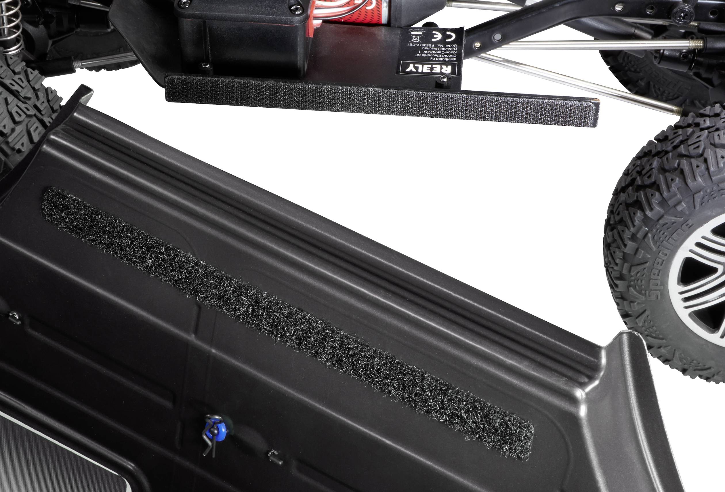 A remote-controlled car in close-up reveals the chassis and tyres. In the background, the gearbox is partially visible.