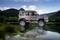 A silver-coloured off-road vehicle is parked on the shore of a lake, surrounded by lush, green trees and wooded hills.