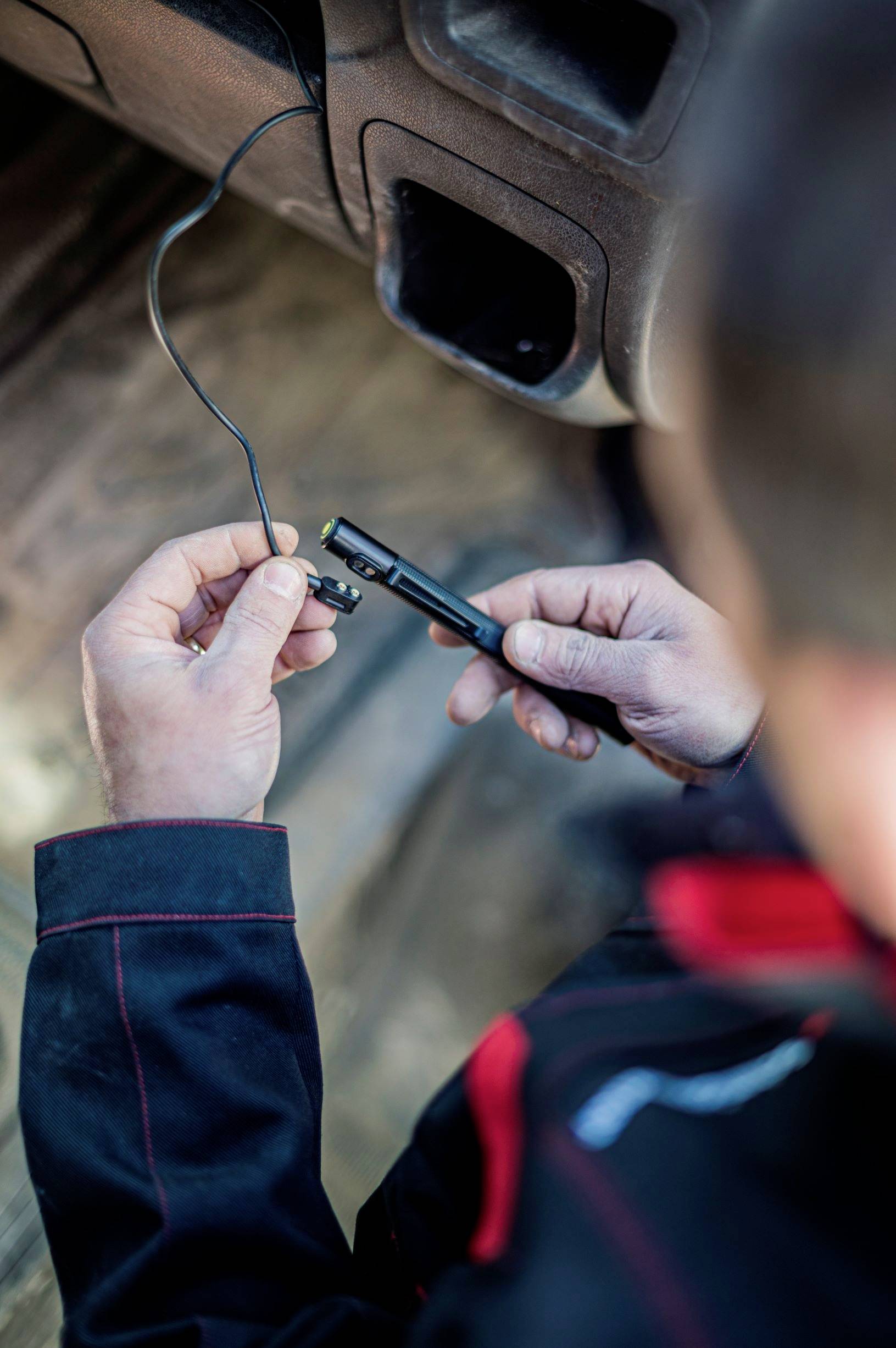 A person in workwear is holding a device with a cable and inspecting it, possibly during a technical repair.