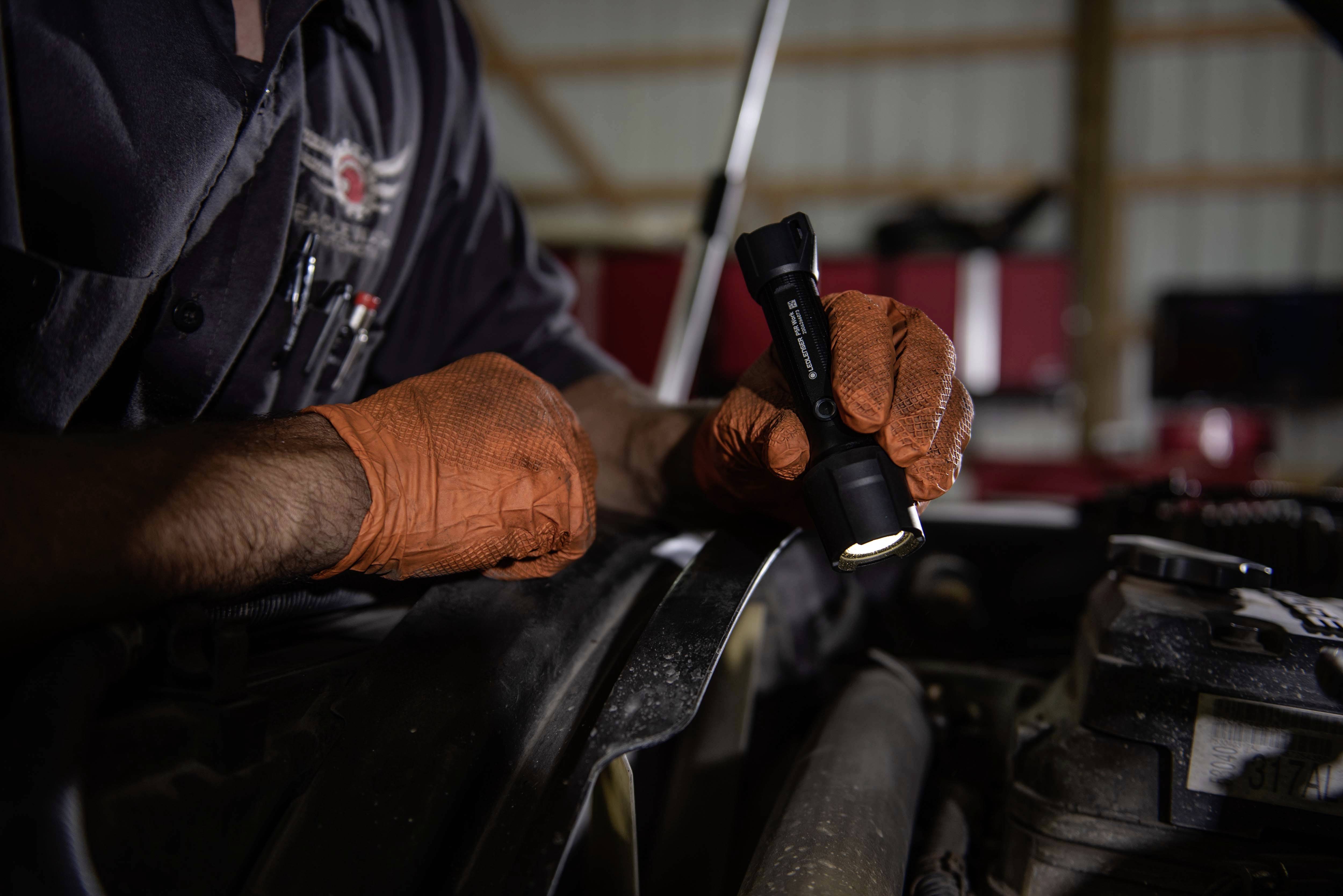 A person wearing orange gloves is holding a torch and inspecting the engine bay of a vehicle in a workshop.
