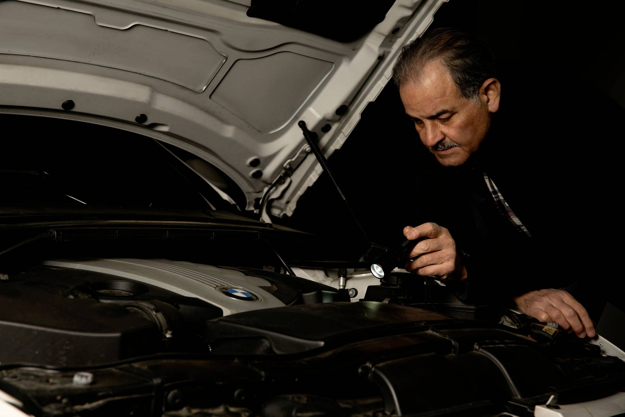 A man is examining the engine of an open car with a torch, focused and precise, in a dark environment.