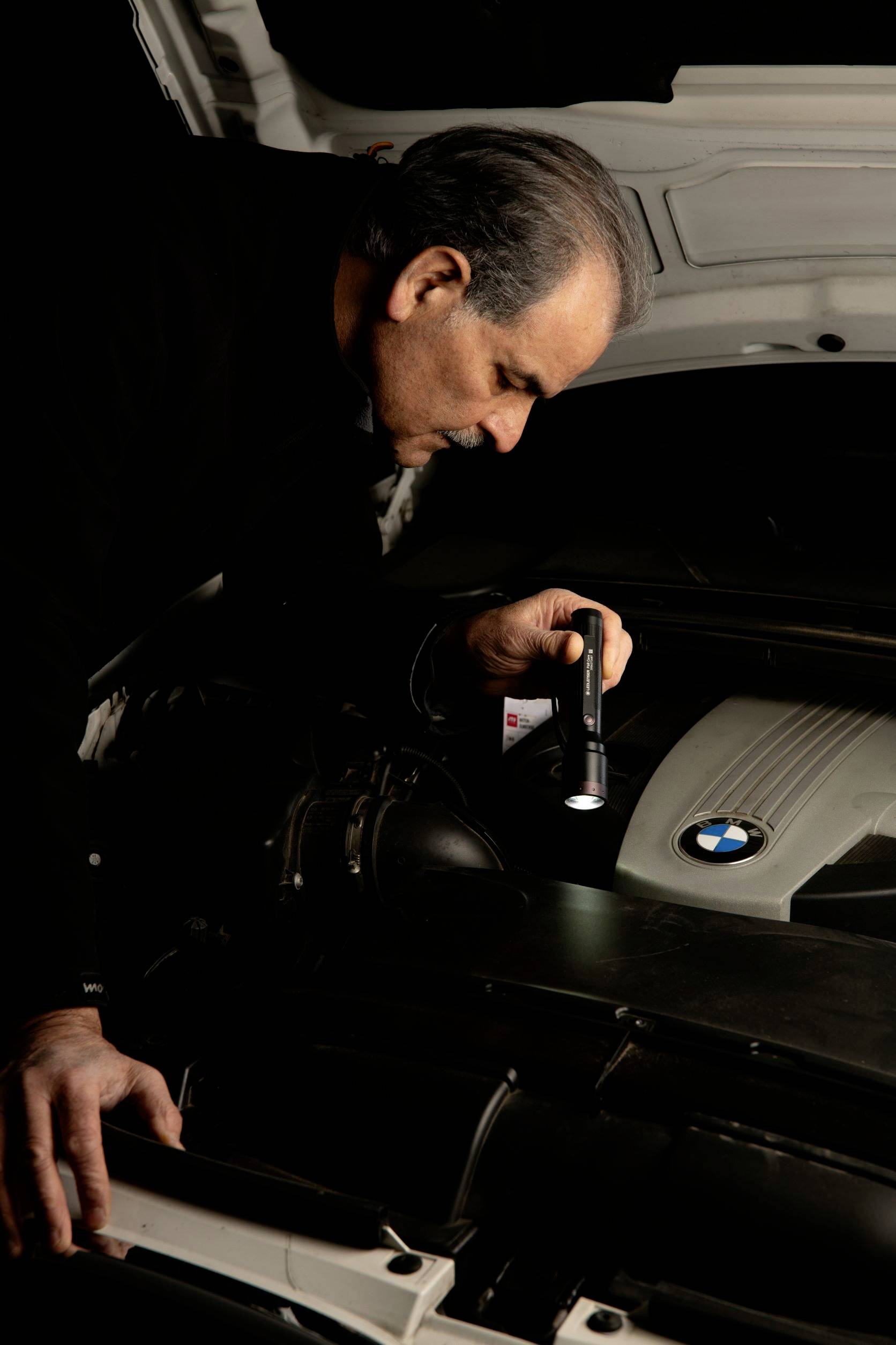 A man is inspecting the engine compartment of a car with a torch. He appears to be searching for a problem or checking something.