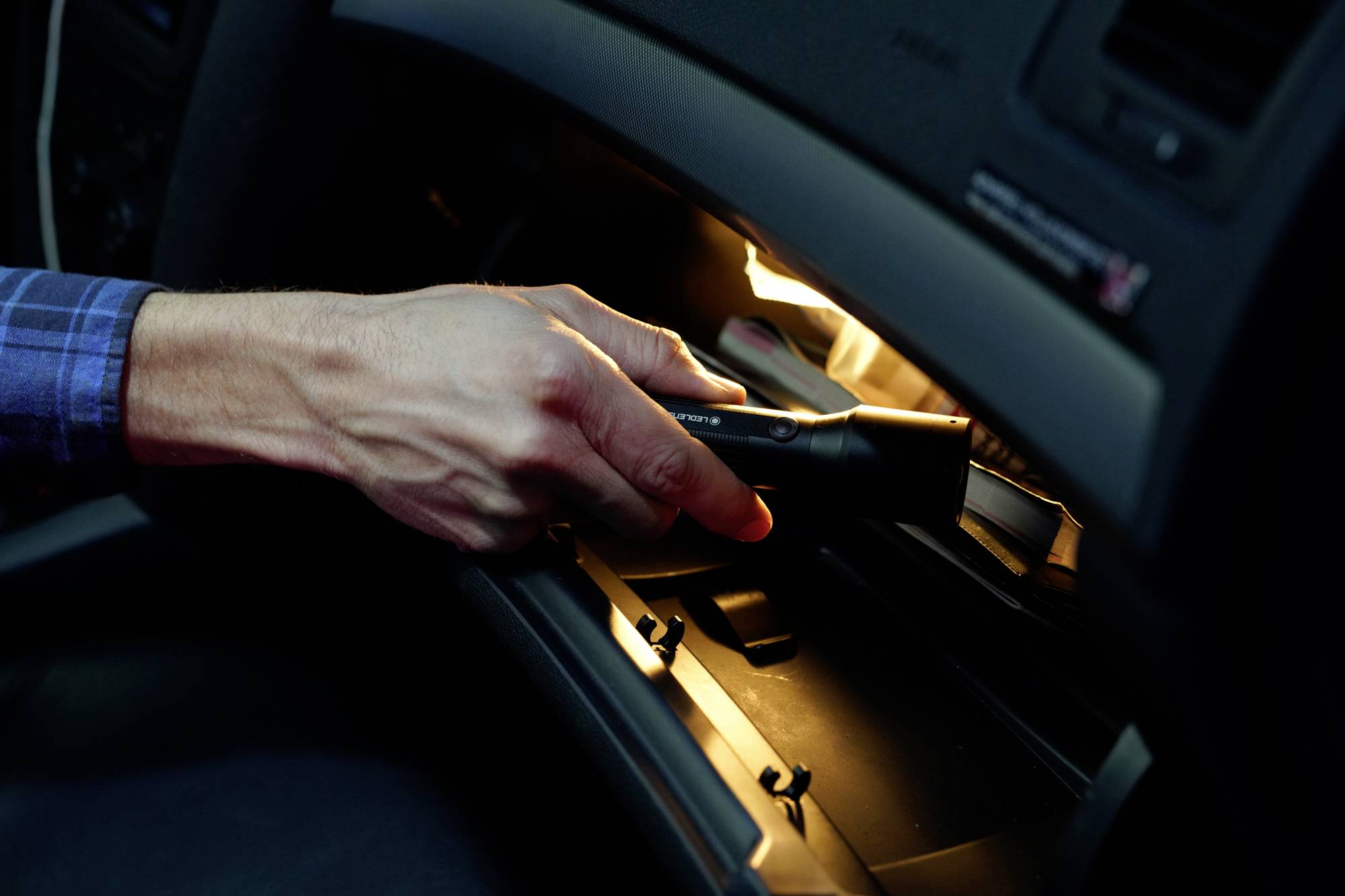 A hand holds a torch and shines it into the open glove compartment of a car.