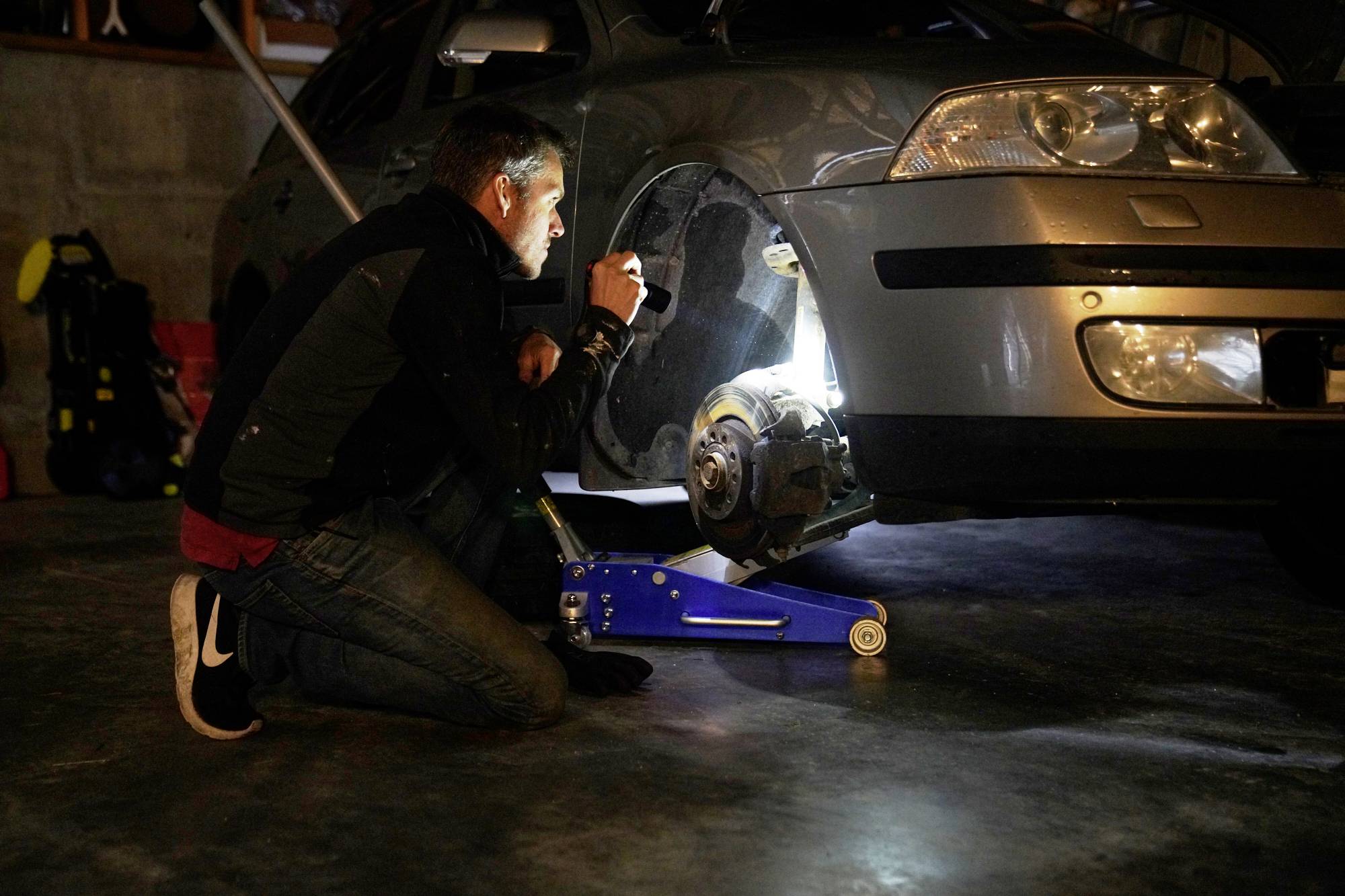 A man is kneeling beside a car raised on a jack and examining the wheel with a torch. The scene is set in a garage.