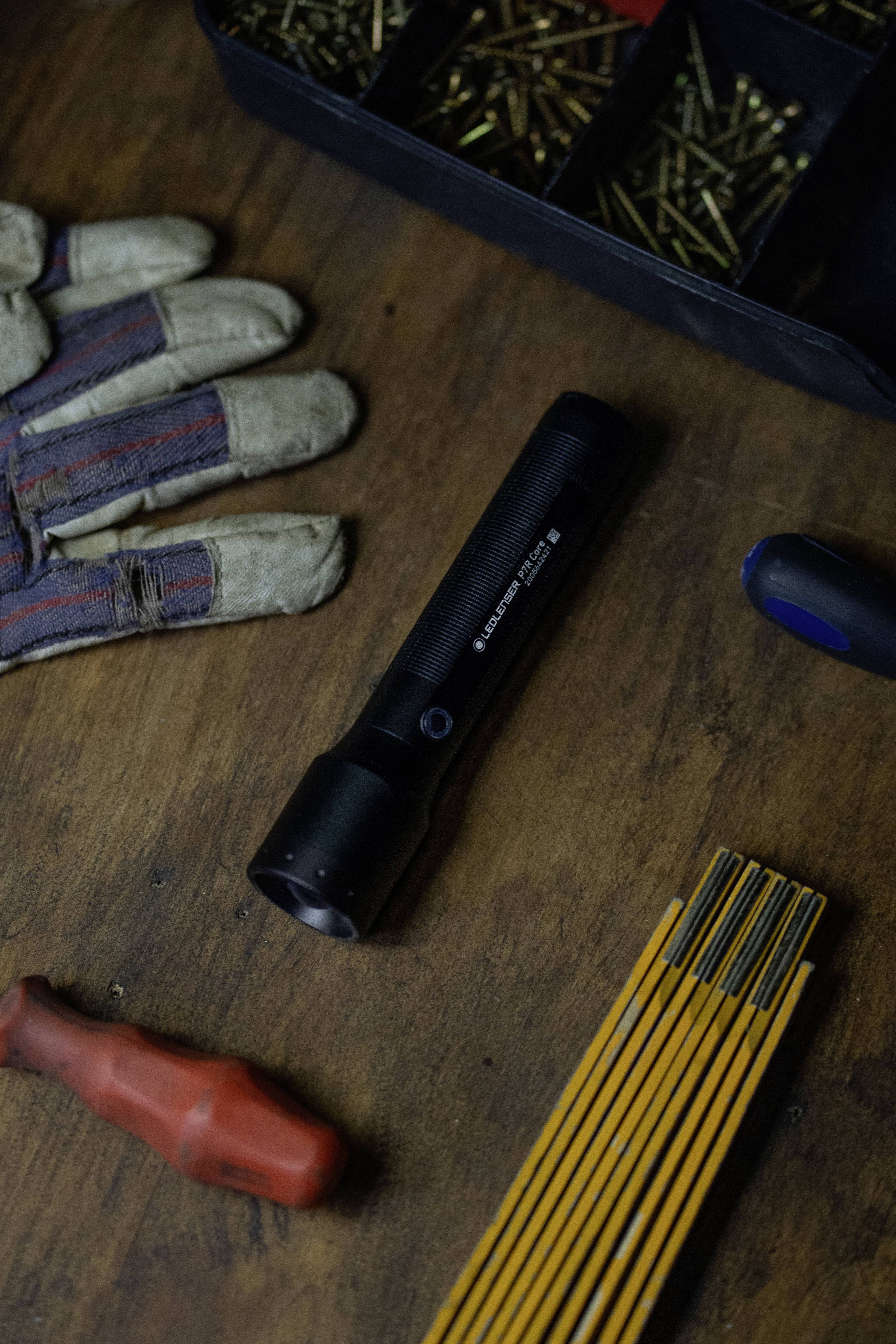 A torch lies on a wooden table, surrounded by screws in a container, a work glove, and tools.