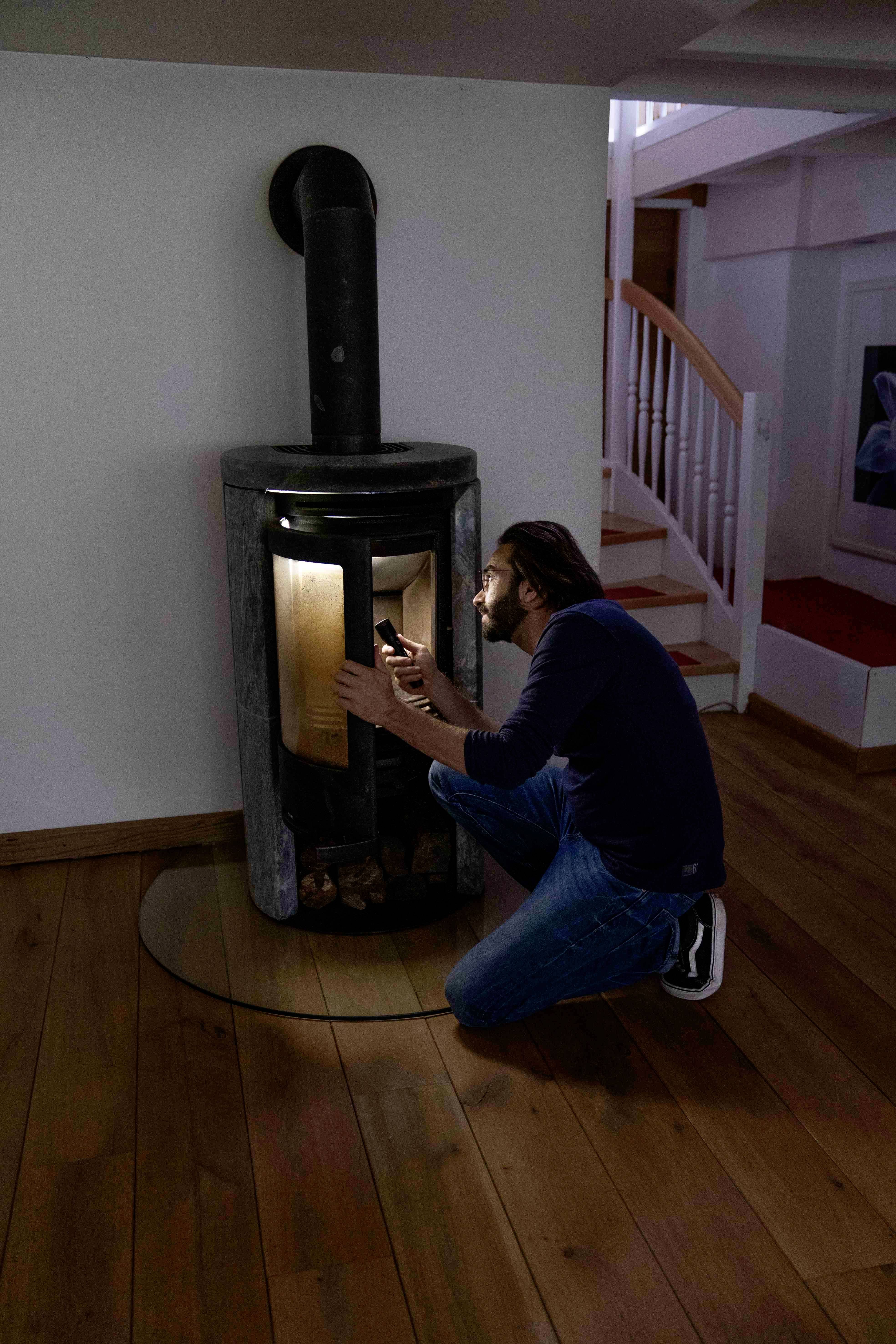 A person is sitting on the floor and lighting a fire in a modern fireplace in a cosy room with a wooden floor.