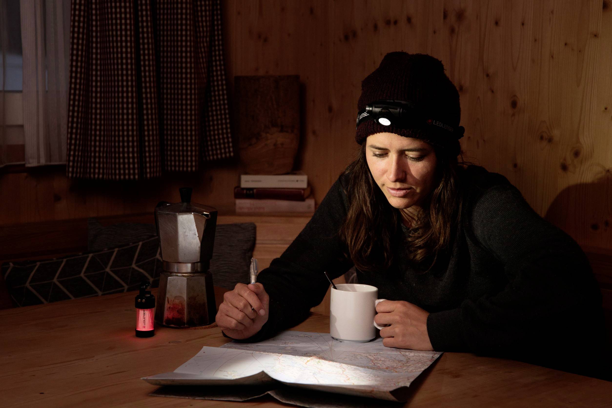 A person is sitting at a wooden table and examining a map. A cup and a coffee pot are placed beside them. The person is wearing a cap with a head torch illuminating the map.