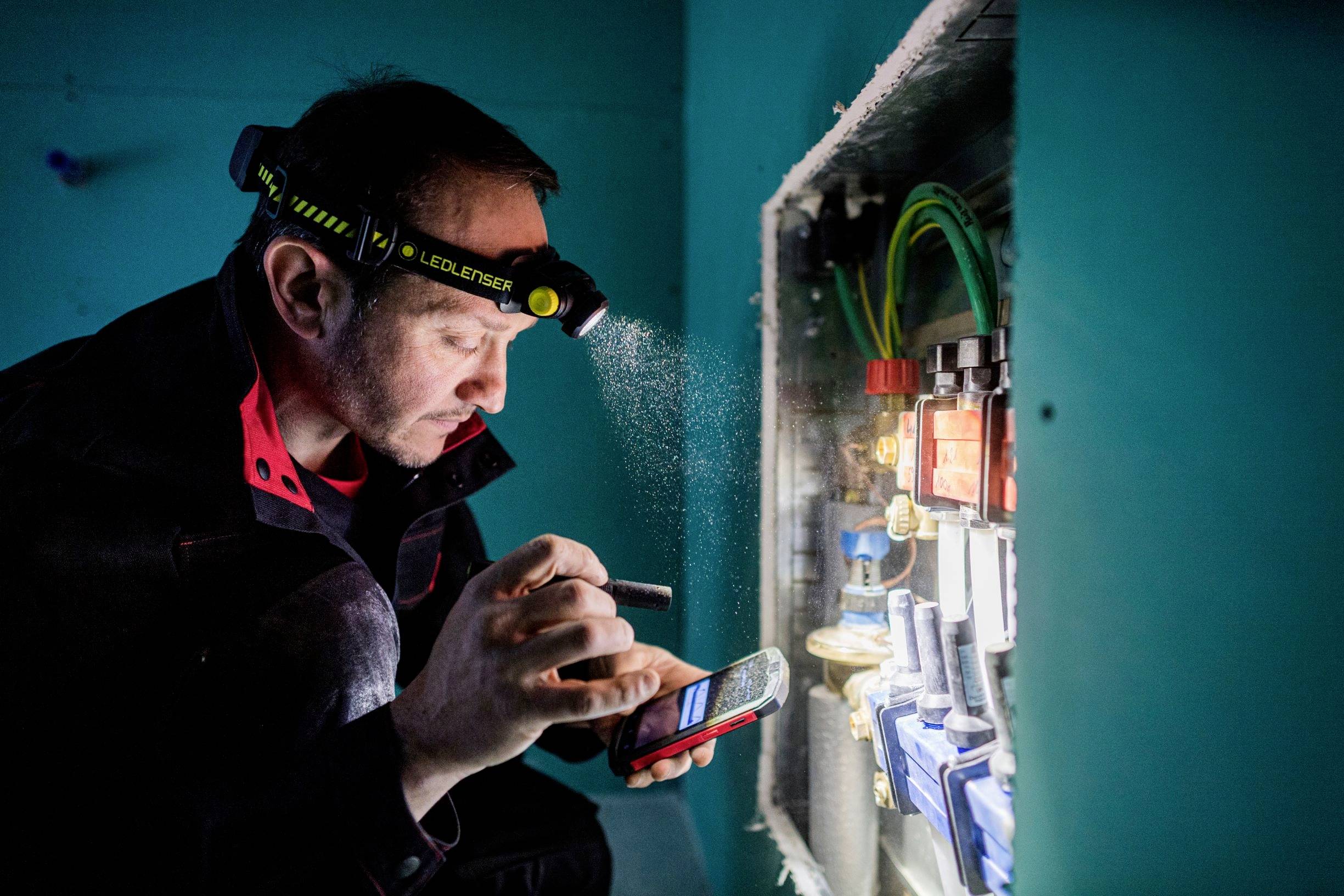 An electrician wearing a head torch is carefully working on a fuse box and checking something on his smartphone.