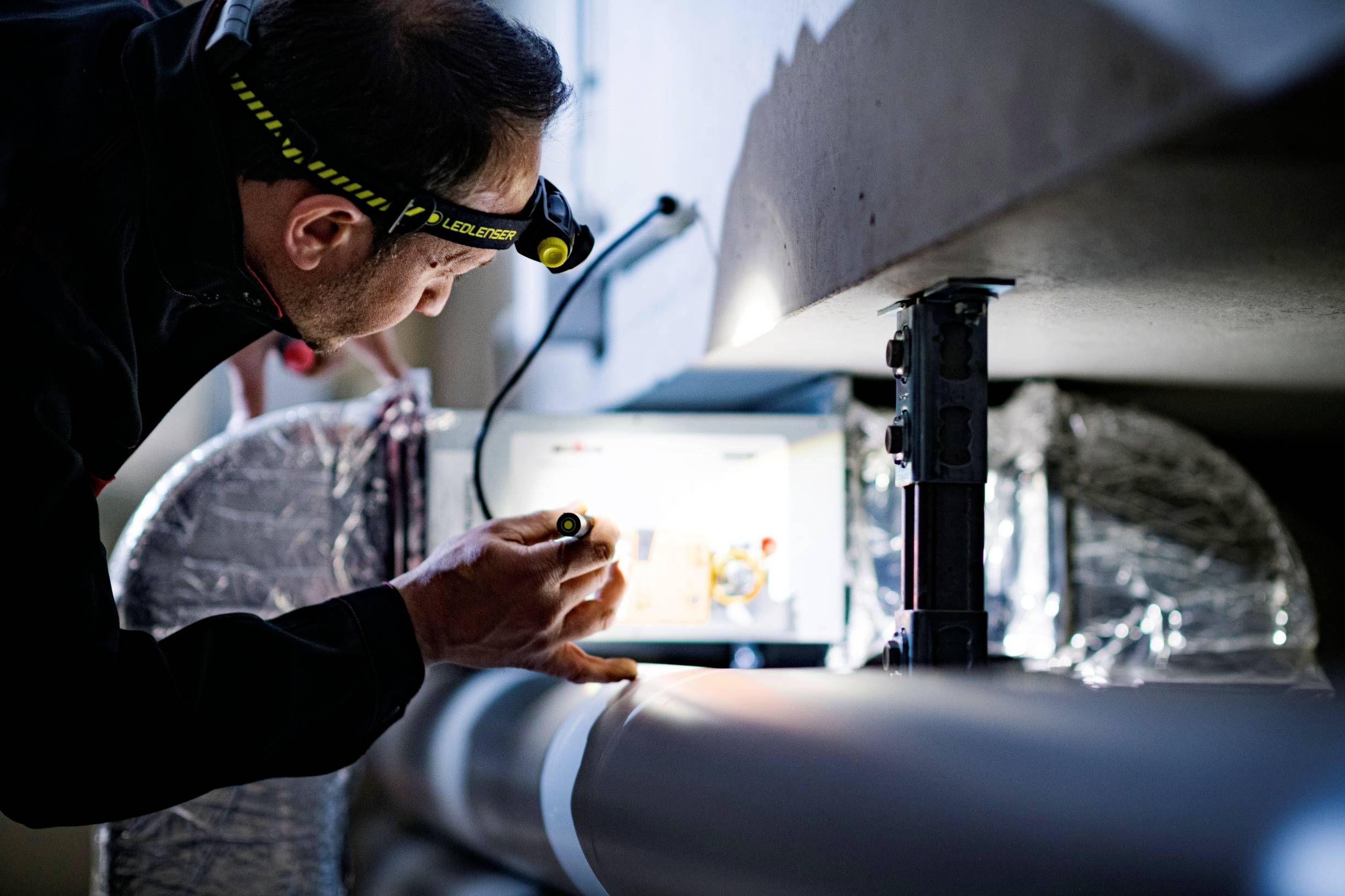 A man wearing a head torch checks technical equipment in a lit room, examining details on a large pipe.