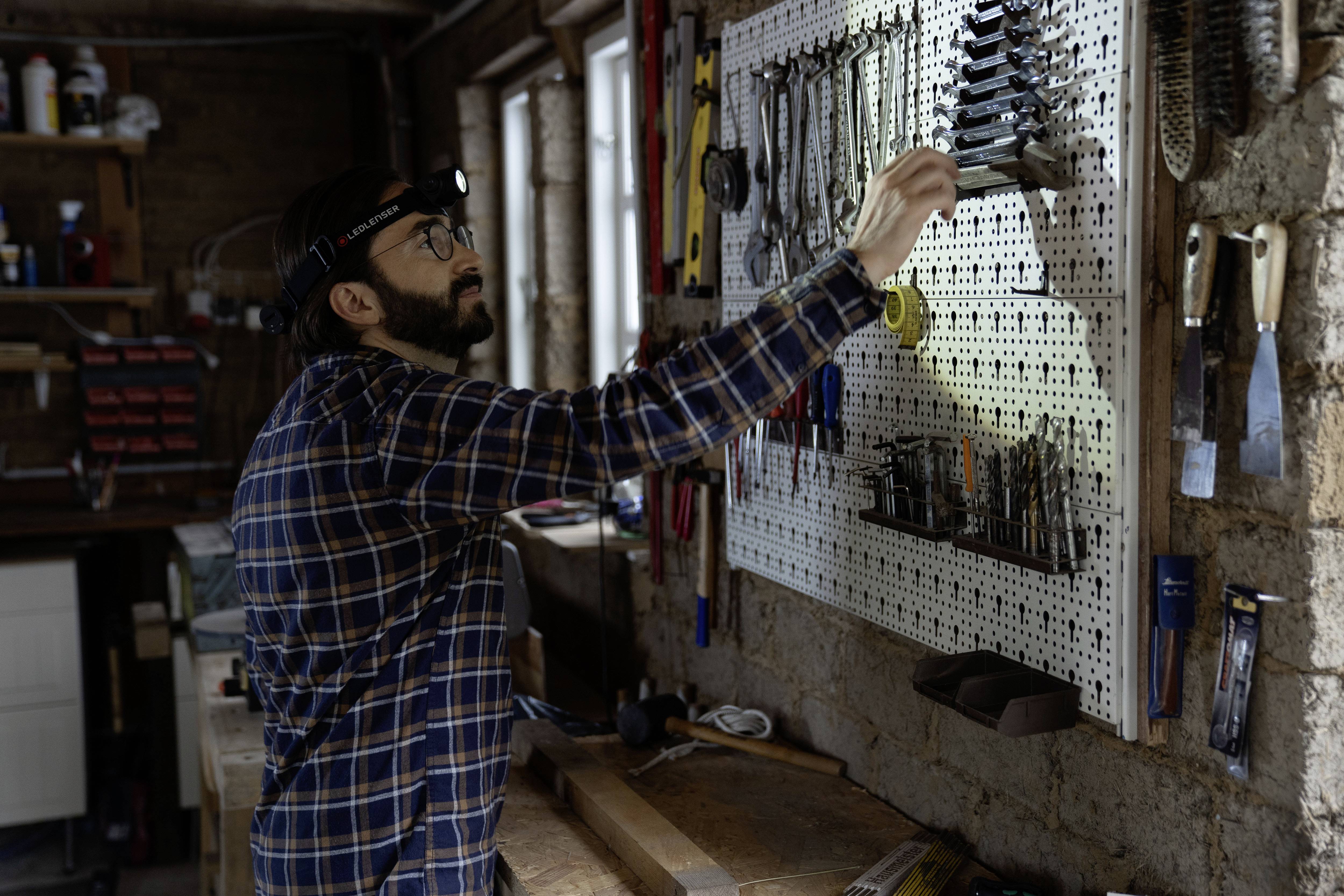 A man wearing a head torch is working in a workshop and searching for tools on a wall-mounted rack.